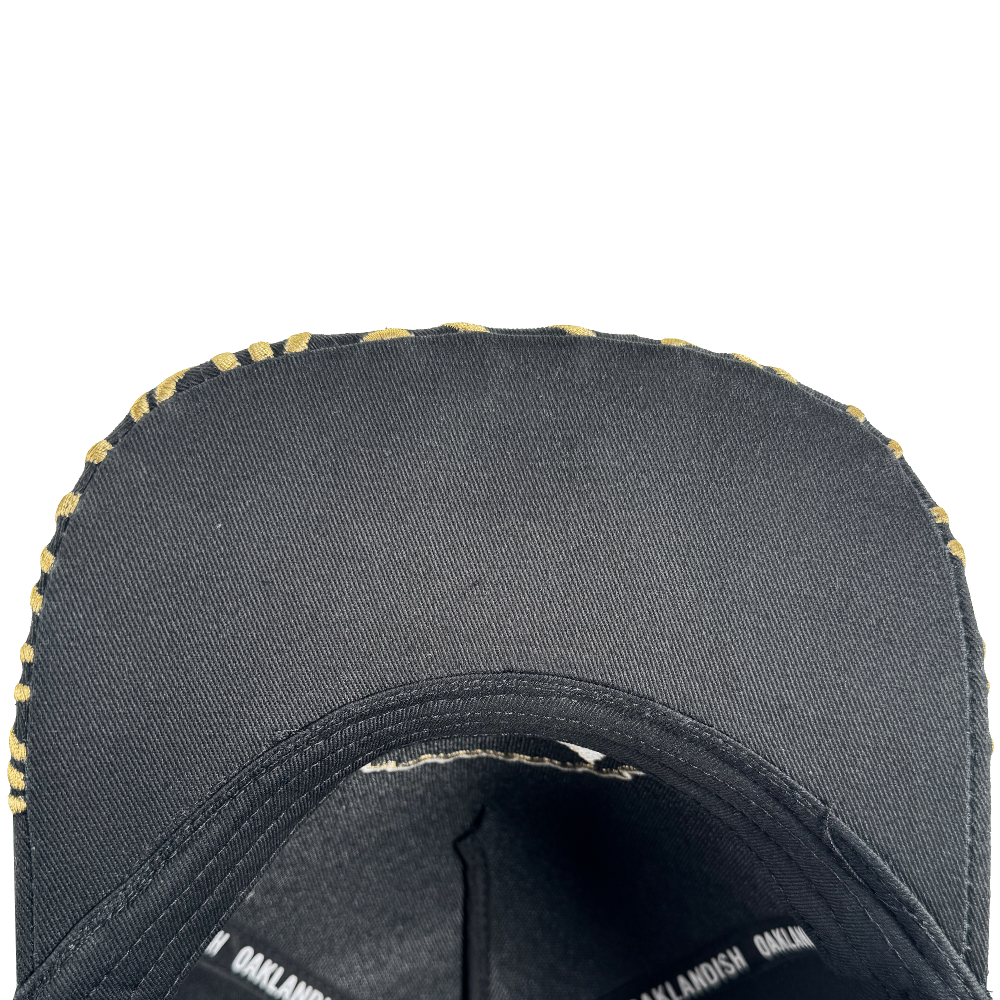 Close-up of the underside of an Oakland Roots SC Growth Cap’s black brim with a yellow camo edge. Inside, black seams and a band with repeating white “OAKLANDISH” text are visible against a white background.