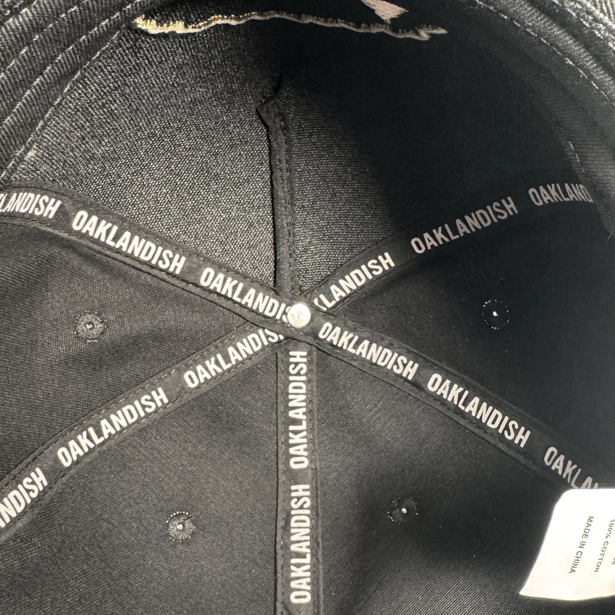 Close-up of the inside of a black Growth Cap by Oakland Roots SC. The inner seams feature black fabric strips displaying "OAKLANDISH" in white, and a white tag is partially visible along the rim under the light.