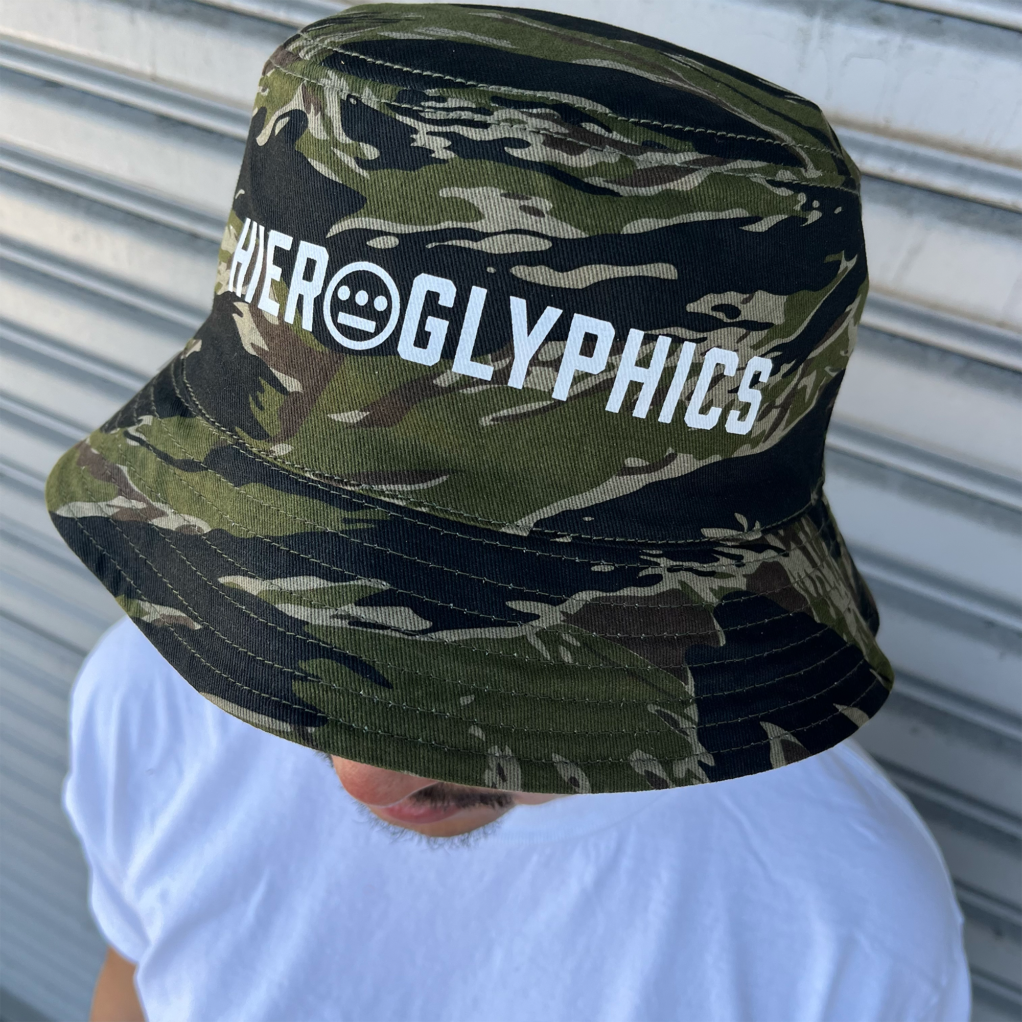 Side view of a man standing outdoors, wearing a green camouflage bucket hat with a white Hieroglyphics Hip Hop Crew logo on the front crown. 