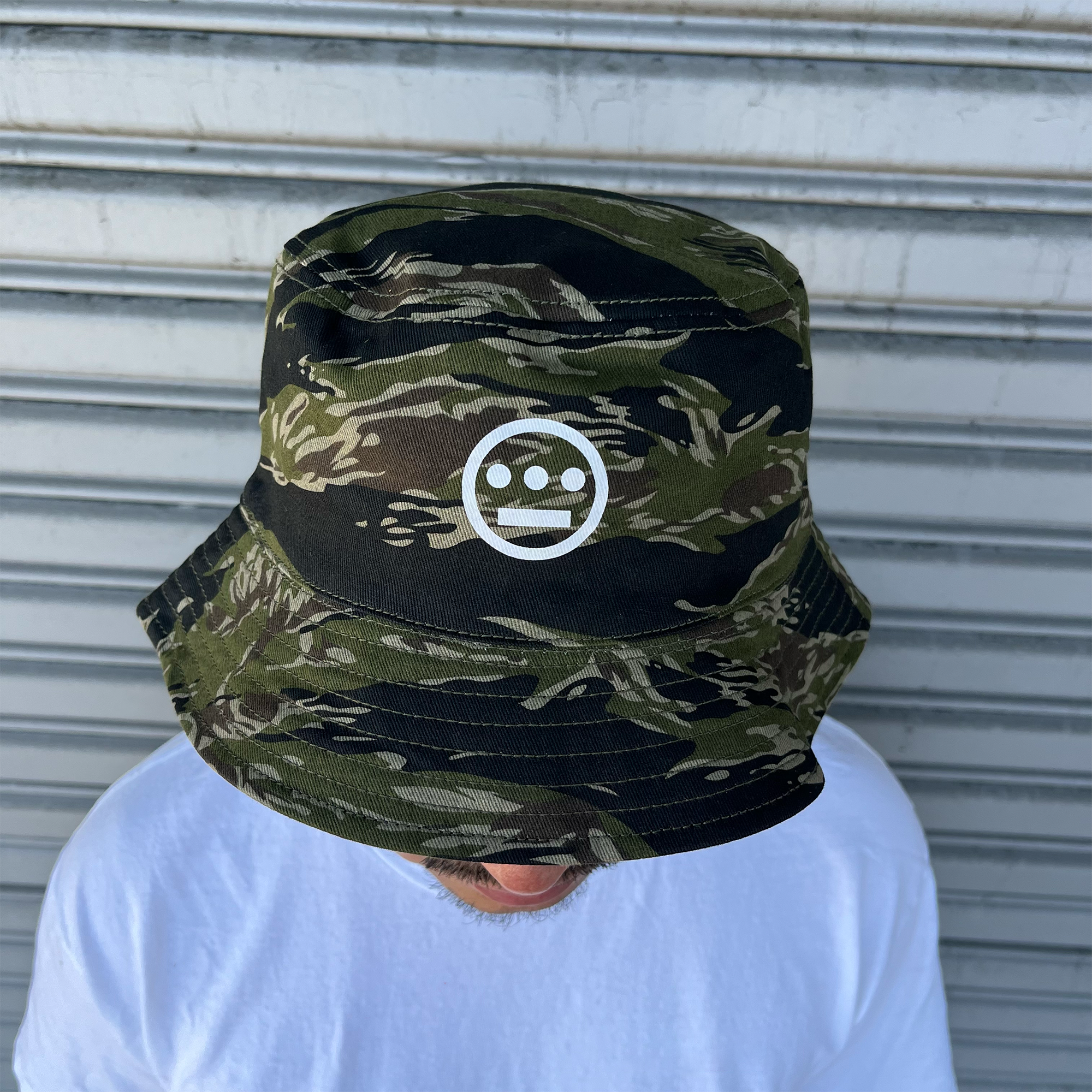 A person is wearing the Hieroglyphics Hiero Reversible Camo Bucket hat, showcasing its dark green tiger camo design and white smiling face logo on the front. The individual, dressed in a plain white T-shirt, is looking downward against a grey, metallic, horizontally ribbed backdrop.