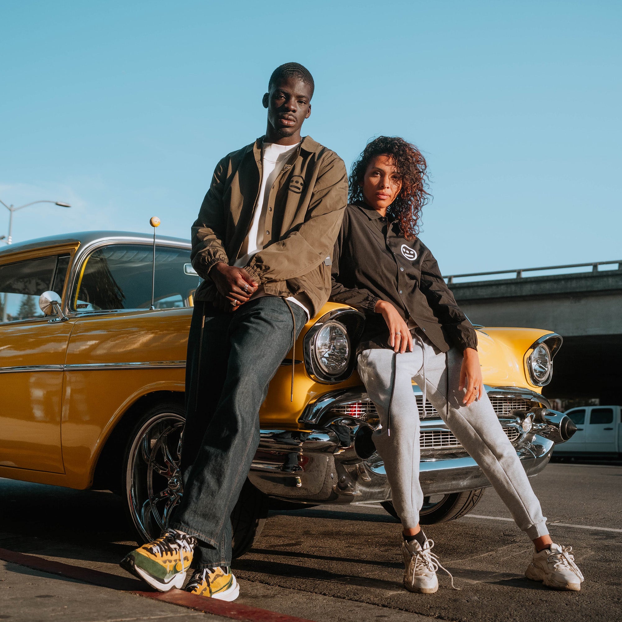 A man and woman leaning against a car, the male wearing an olive jacket with a black Hiero logo and the female wearing a black jacket with a white Hiero logo.