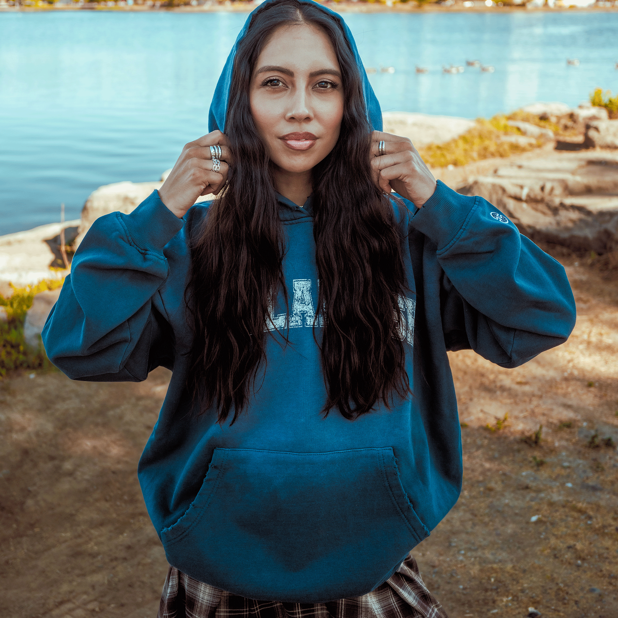 A young woman with long, wavy dark hair stands by a lake, calmly looking at the camera while wearing the Oaklandish Homage Hoodie with the hood up and plaid pants. She holds the drawstrings; ducks and rocks appear in the background.