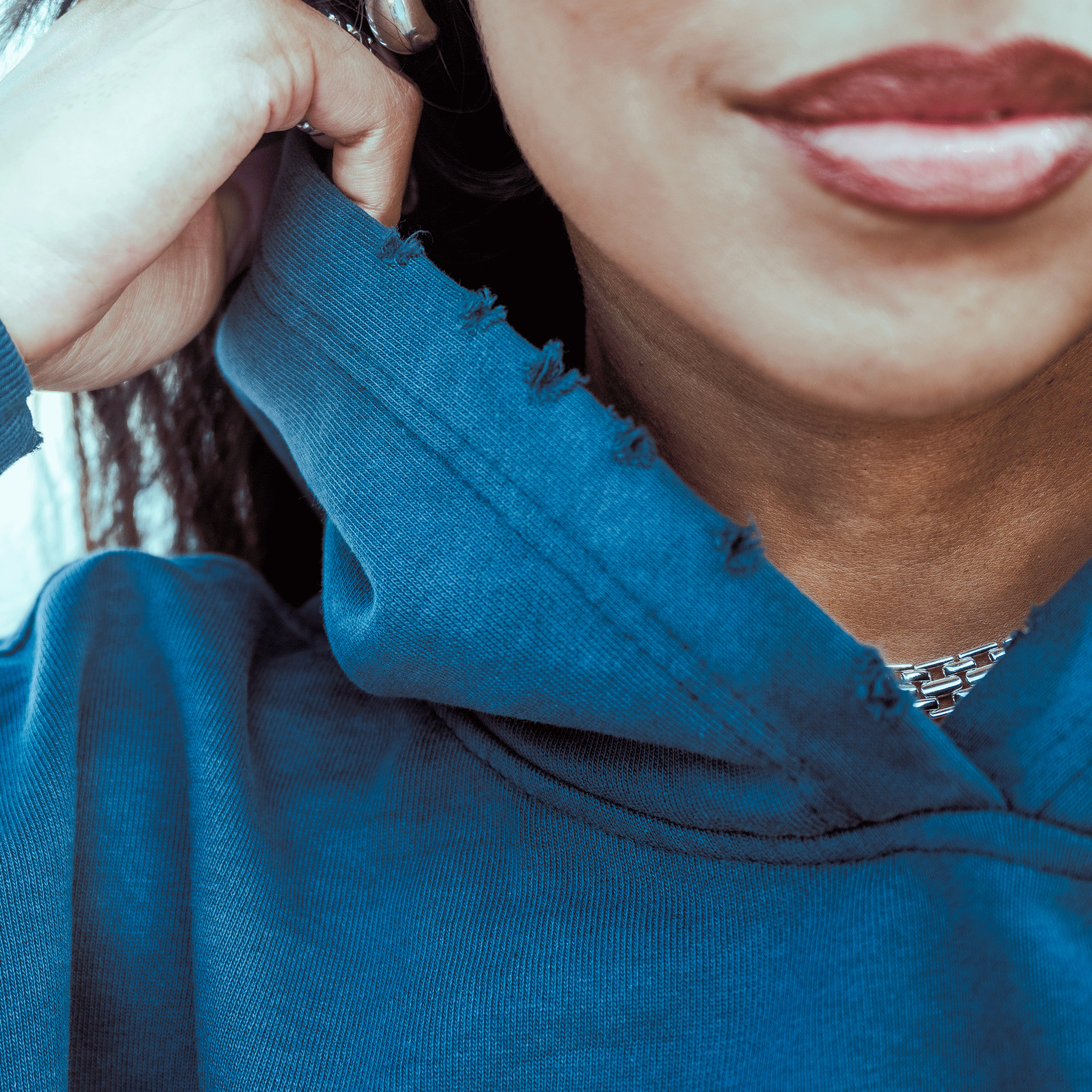 A close-up of a person with medium brown skin wearing bright red lipstick, a silver-link necklace, and the Oaklandish Homage Hoodie with a frayed collar. They adjust the oversized pullover hoodie with one hand.