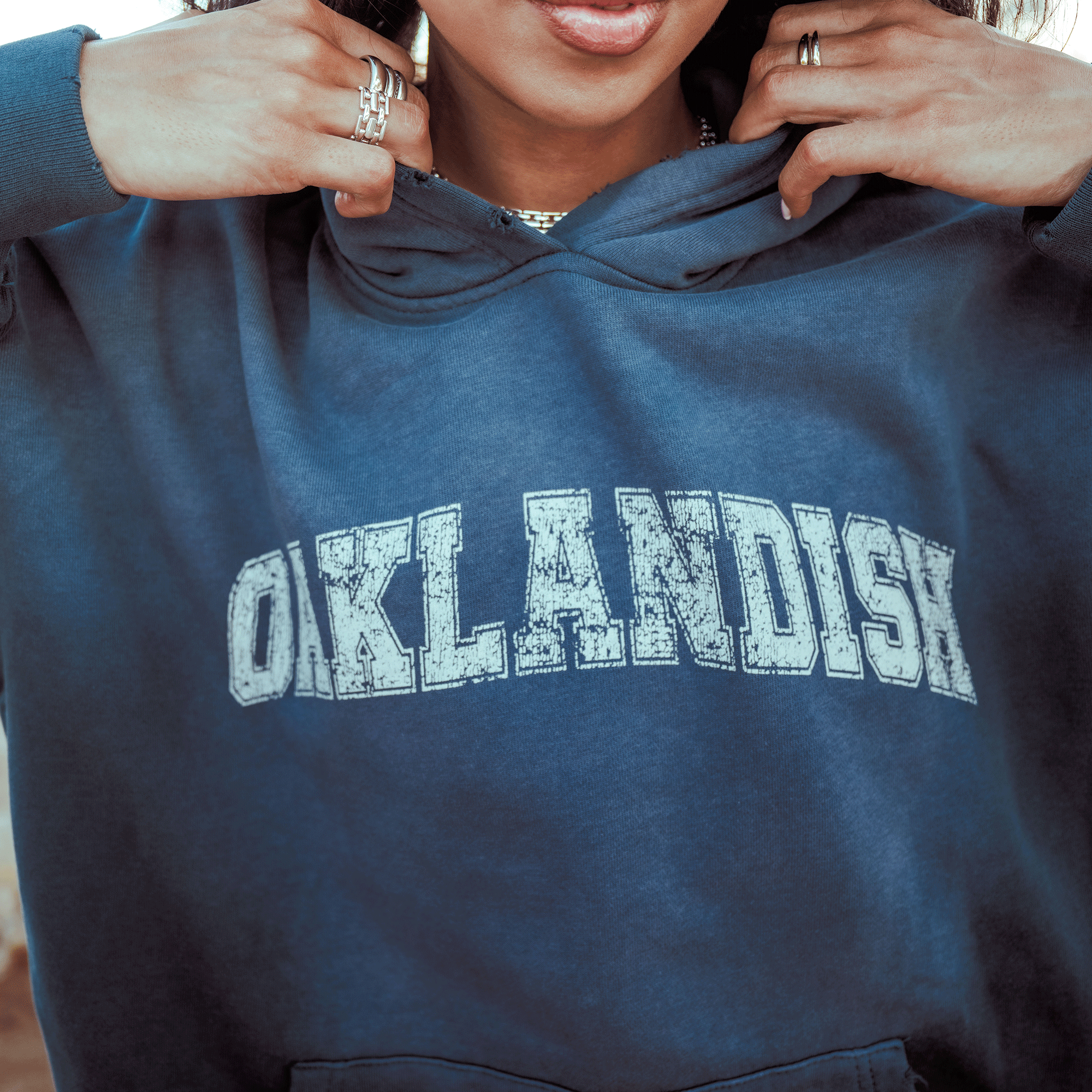 A person tugs at the collar of an oversized blue Oaklandish Homage Hoodie, showing off bold, distressed "OAKLANDISH" lettering across the chest. Only their hands with rings and the lower half of their face are visible.