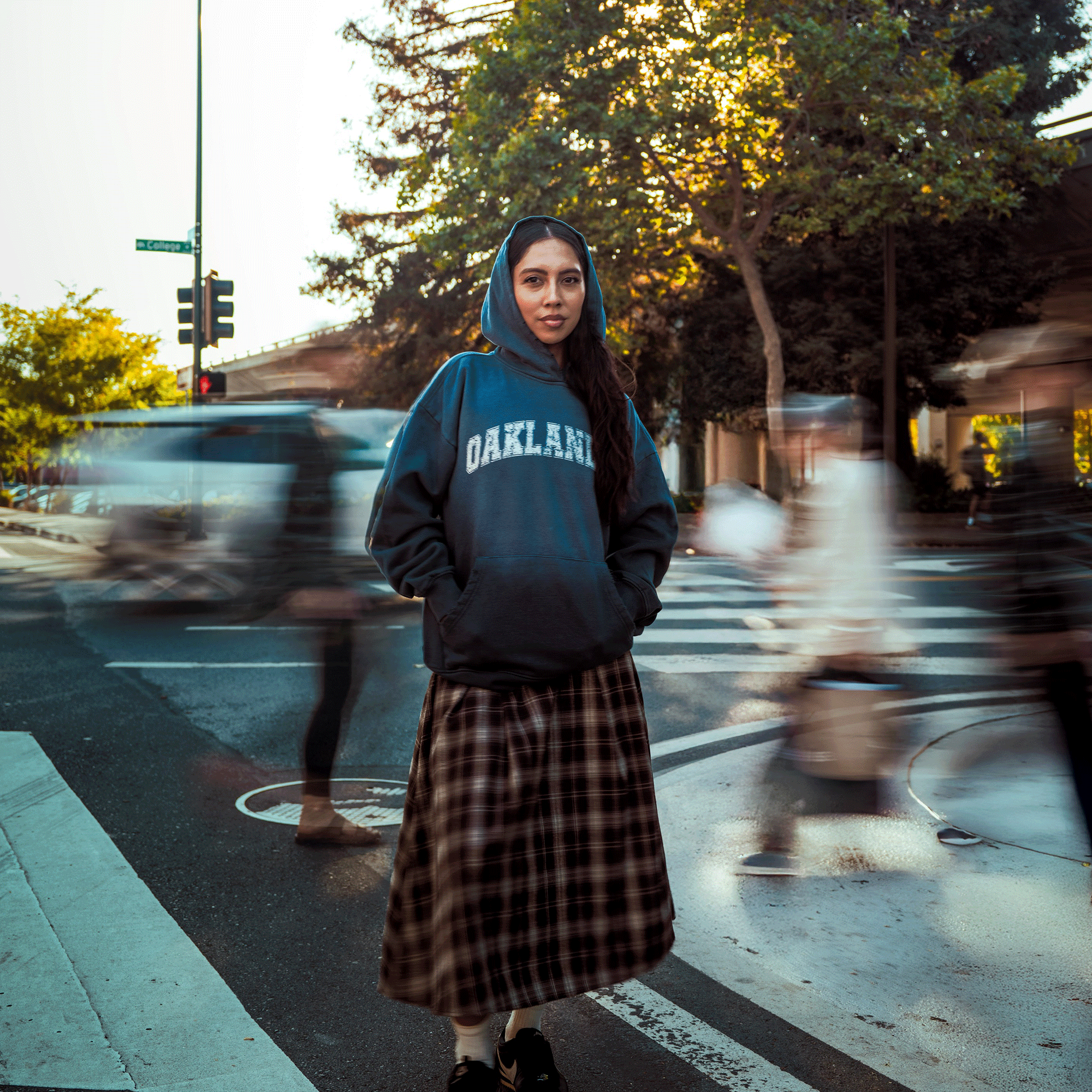 A woman stands confidently on a city street corner in an Oaklandish Homage Hoodie, blue and oversized with “OAKLAND,” paired with a plaid skirt. Hands in pockets, she faces the camera as blurred pedestrians pass and sunlight streams through trees behind her.