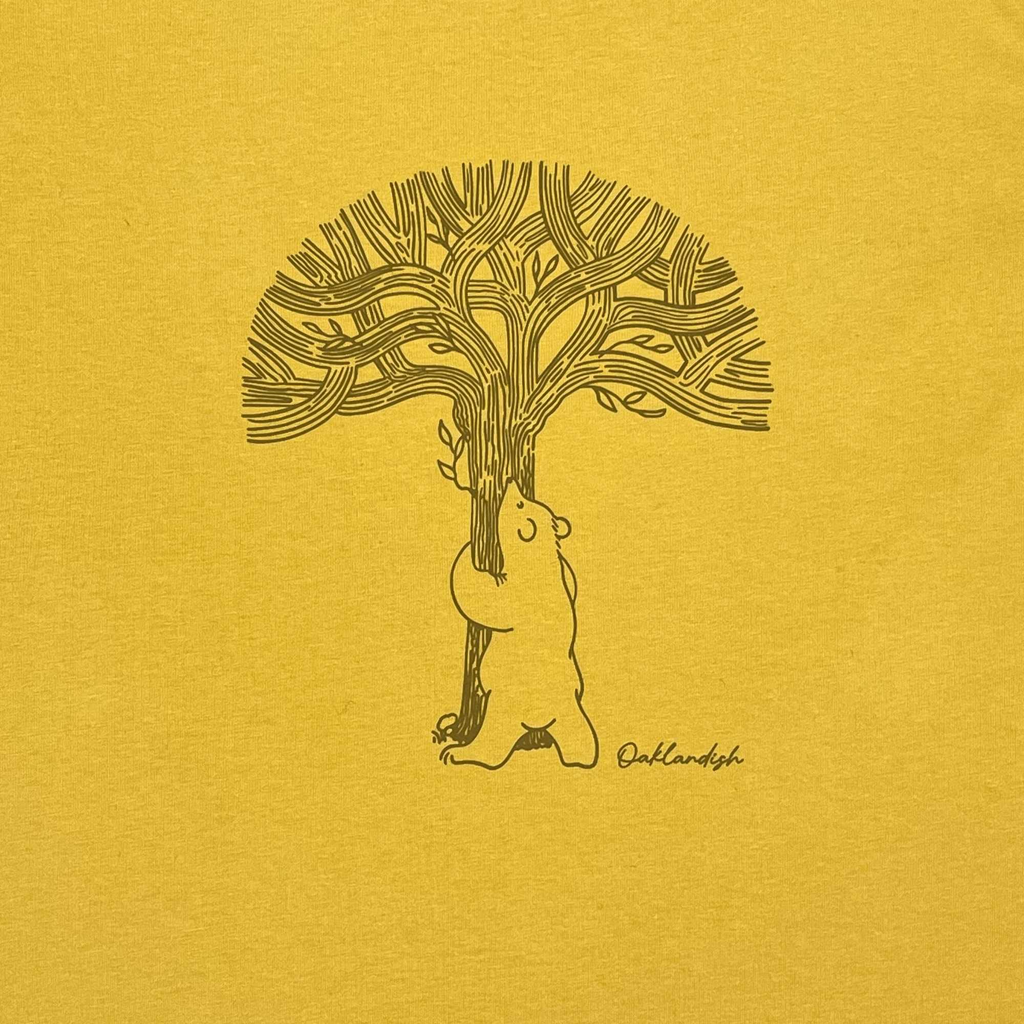 Close-up of line graphic featuring a bear hugging a tree representing Oaklandish with Oaklandish wordmark on mustard yellow women’s cut t-shirt.