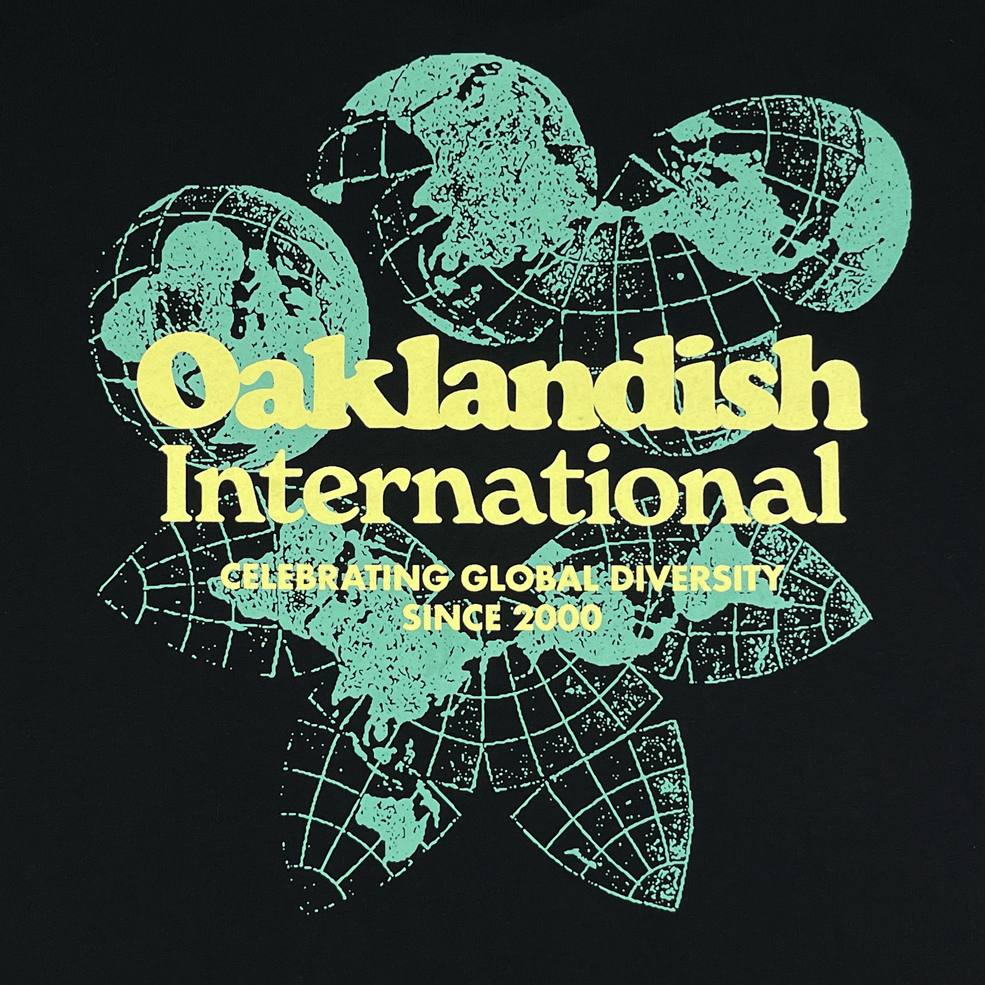 Close-up of green and yellow Oakland International graphic with a caption celebrating global diversity since 2000 on a black tee.