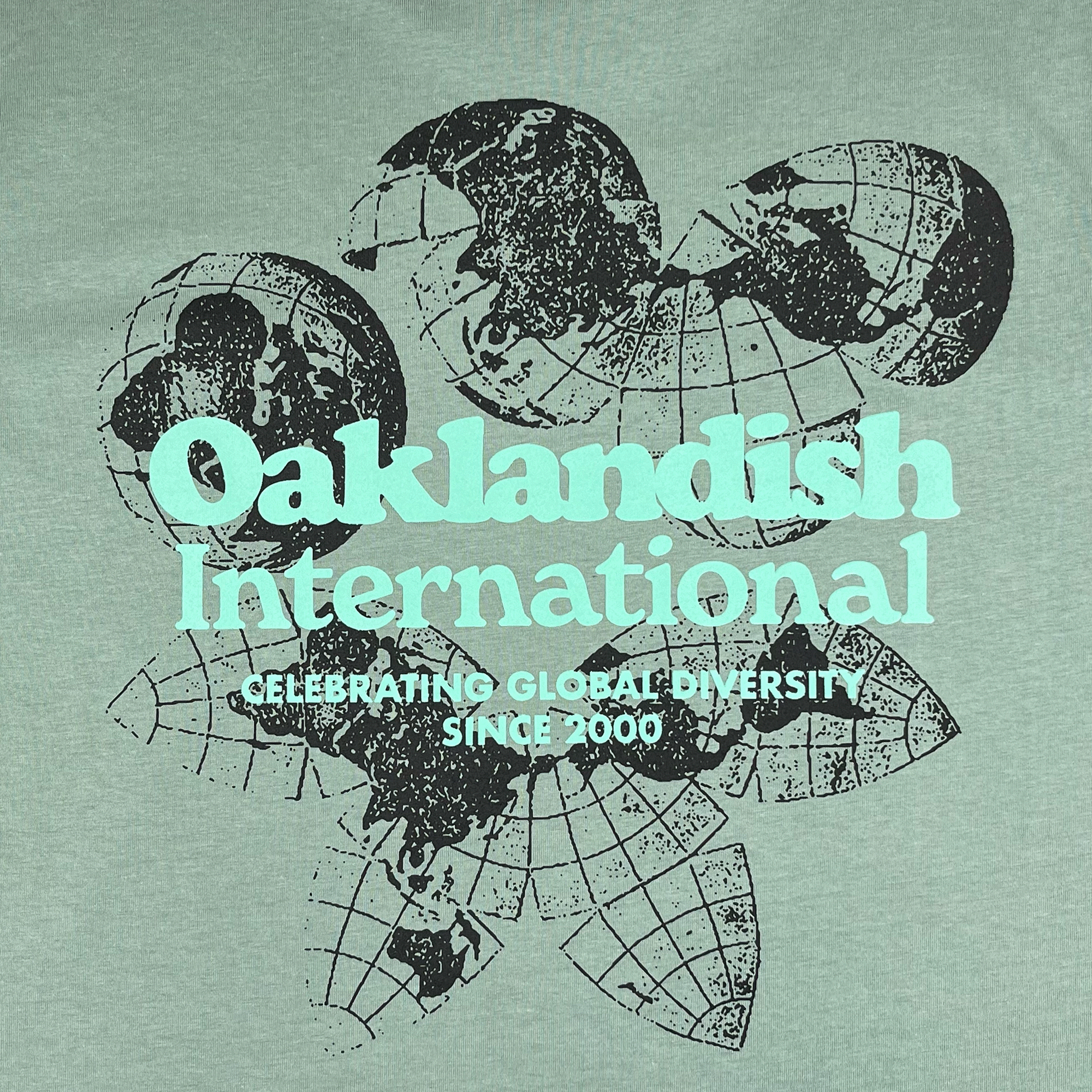 Close-up of Oakland International graphic with a caption celebrating global diversity since 2000 on a sage green tee.