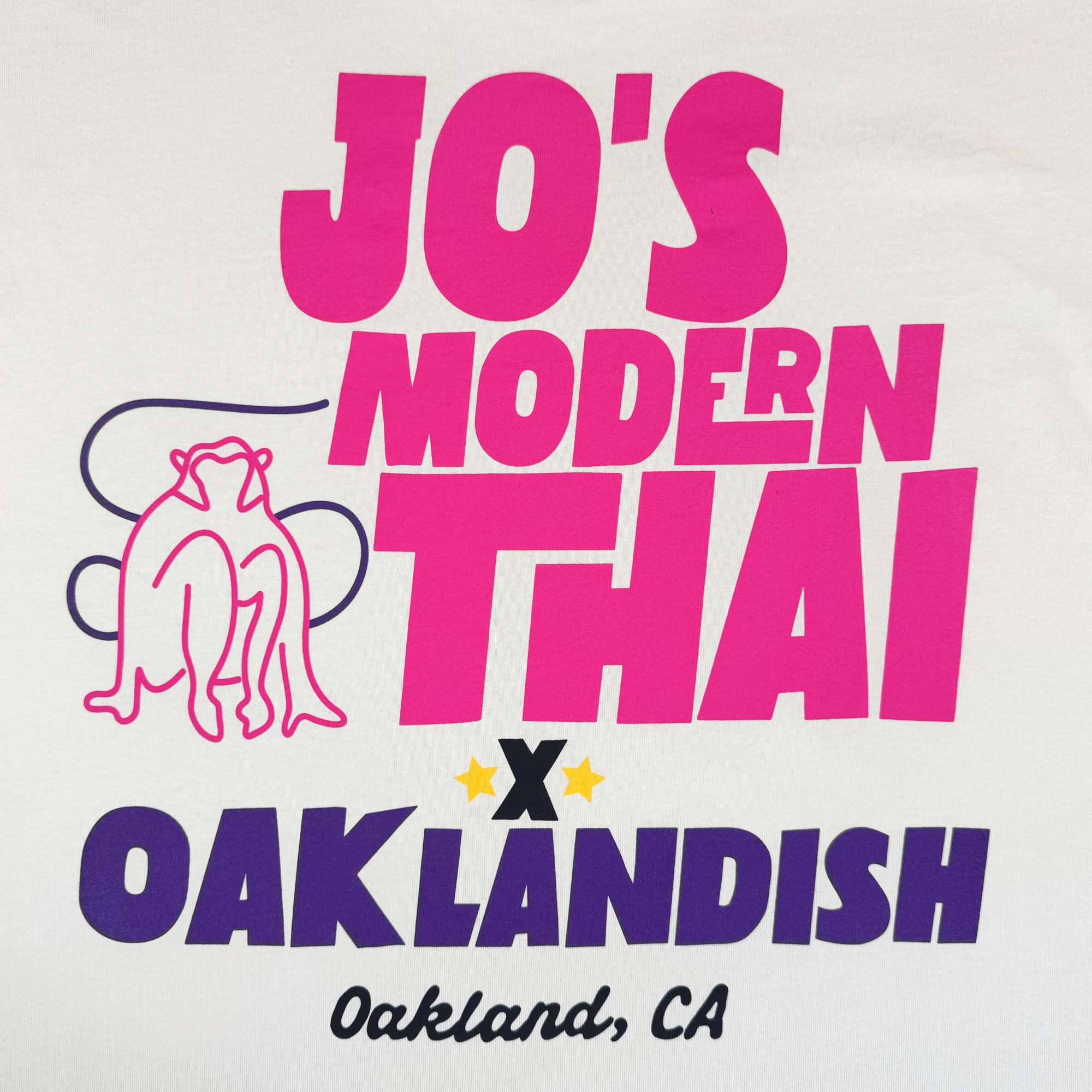 The Jo's Modern Thai X Oaklandish Tee by Oaklandish features bold pink and purple text, “Oakland, CA” in cursive, a line-drawn frog on the left, and two yellow stars above “OAKLANDISH,” all on a heavy-weight t-shirt.