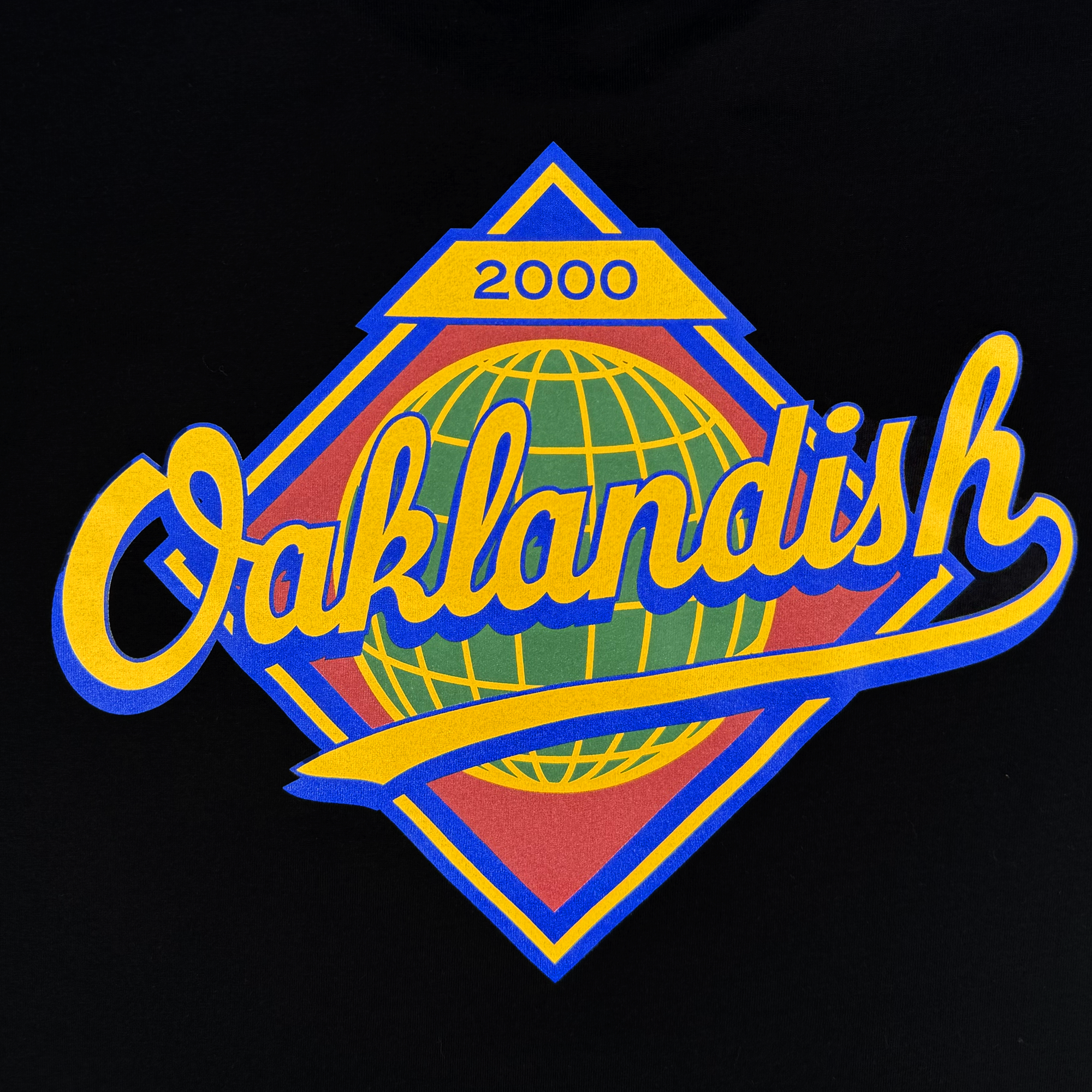 The Leadoff Tee by Oaklandish displays "Oaklandish" in cursive yellow with a blue outline over a vibrant globe on a black t-shirt. Made from 100% cotton, it comes in men's sizes and features a unique yellow diamond with "2000" above the design.