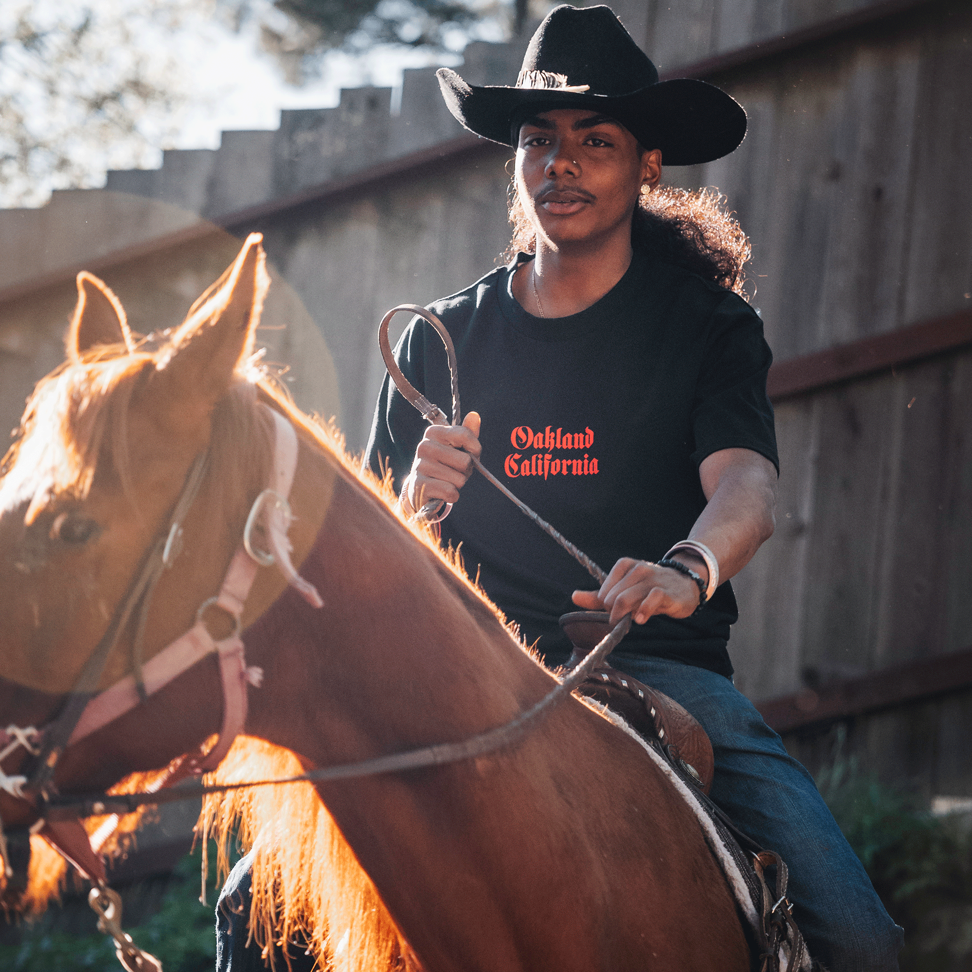 Model on a horse wearing  black tee with Oakland California printed centered in red ink.