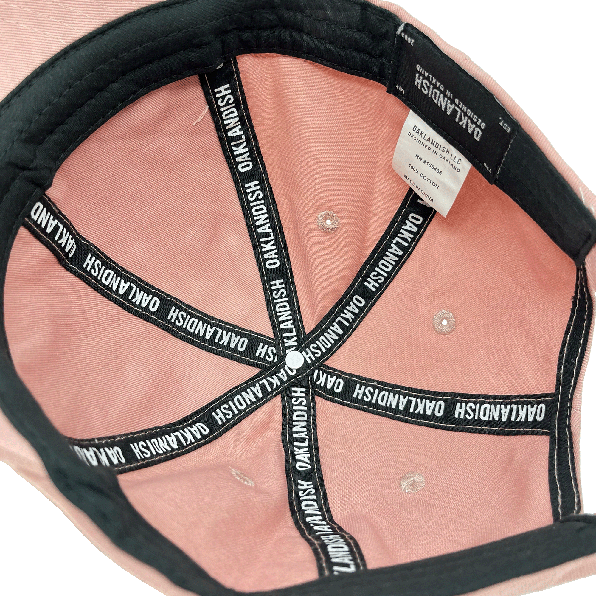 The image displays the interior of an Oaklandish Micro Logo Dad Hat in pink. The black fabric bands within the cap are adorned with the word "OAKLANDISH" repeatedly printed in white. A white and grey label featuring care instructions is stitched beneath the rim, revealing the interior lining. The adjustable strap at the back is also visible.