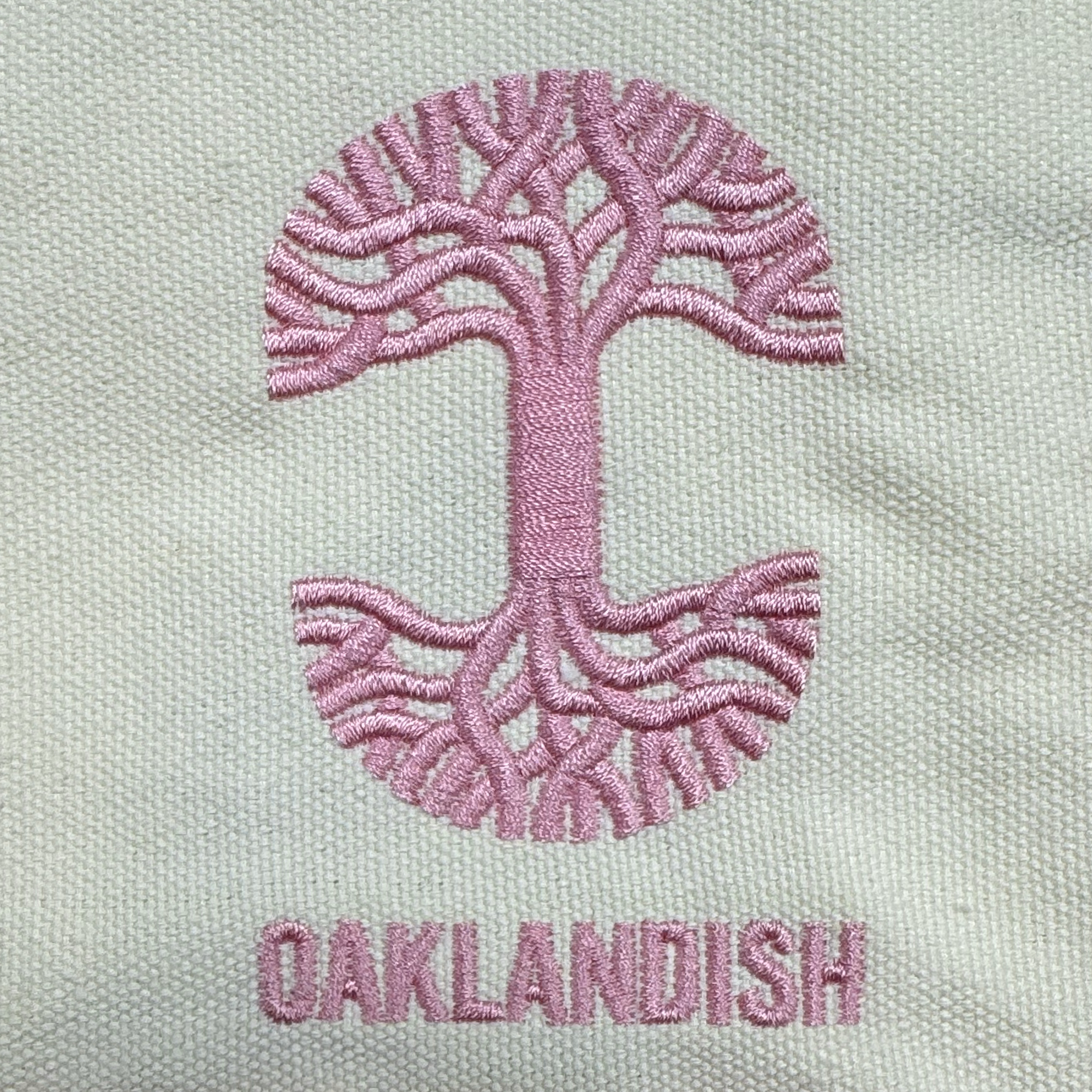 A pink stitched emblem of a stylized tree forms an “O” on the Oaklandish Mini Grocery Tote. Below, “OAKLANDISH” is embroidered in matching pink thread on light fabric. Brand: Oaklandish.