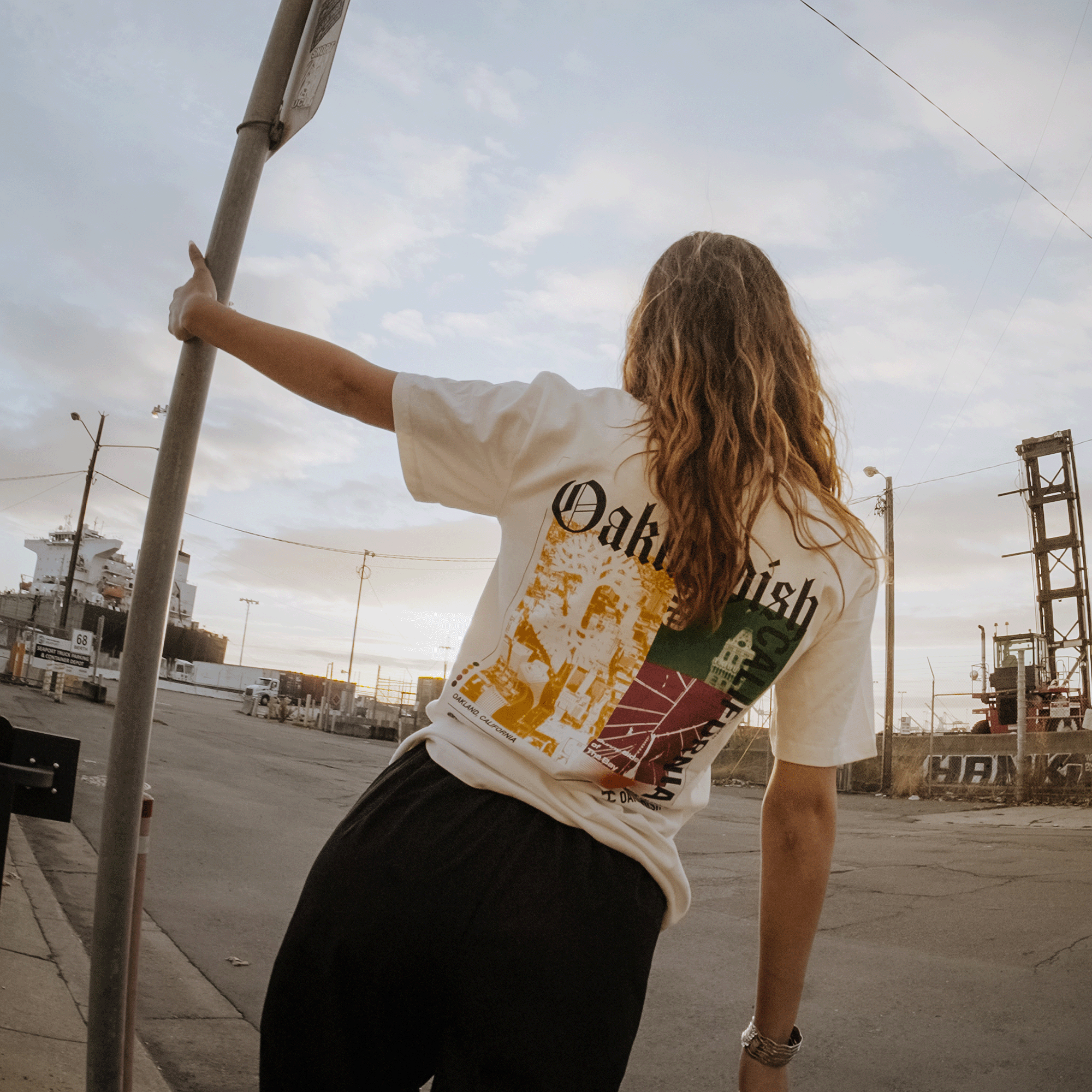Model showing backside view of a natural cotton-colored t-shirt with an abstract yellow, green, and red motif graphic with Oaklandish California wordmarks and logos.