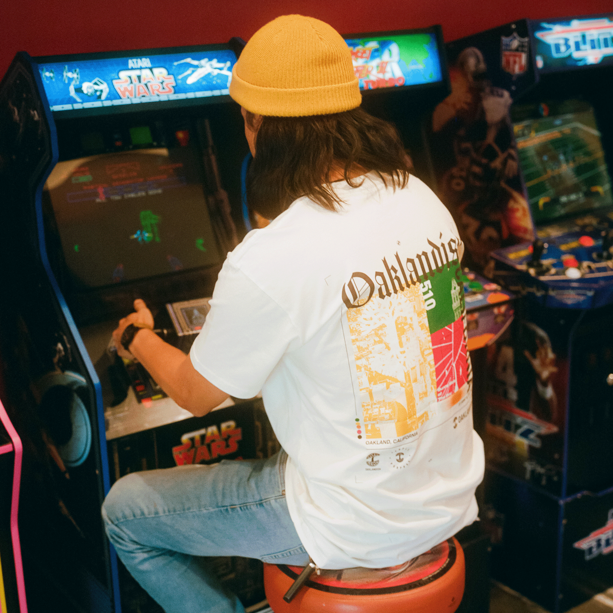 Backside view of a model wearing natural cotton-colored t-shirt with an abstract yellow, green, and red motif graphic with Oaklandish California wordmarks and logos while playing video games.