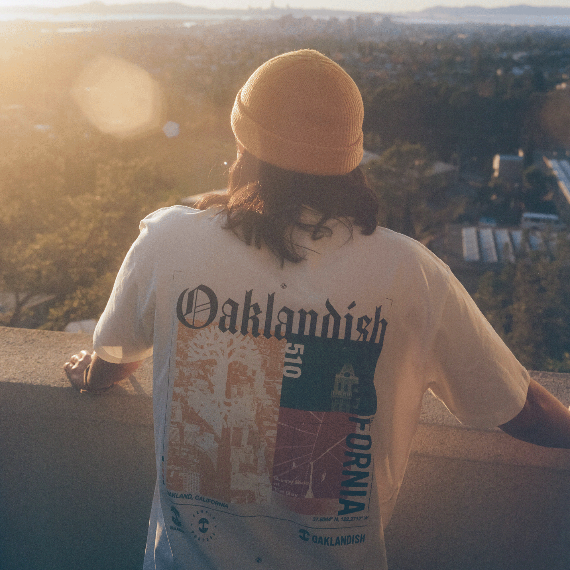 Backside view of a model wearing natural cotton-colored t-shirt with an abstract yellow, green, and red motif graphic with Oaklandish California wordmarks and logos.