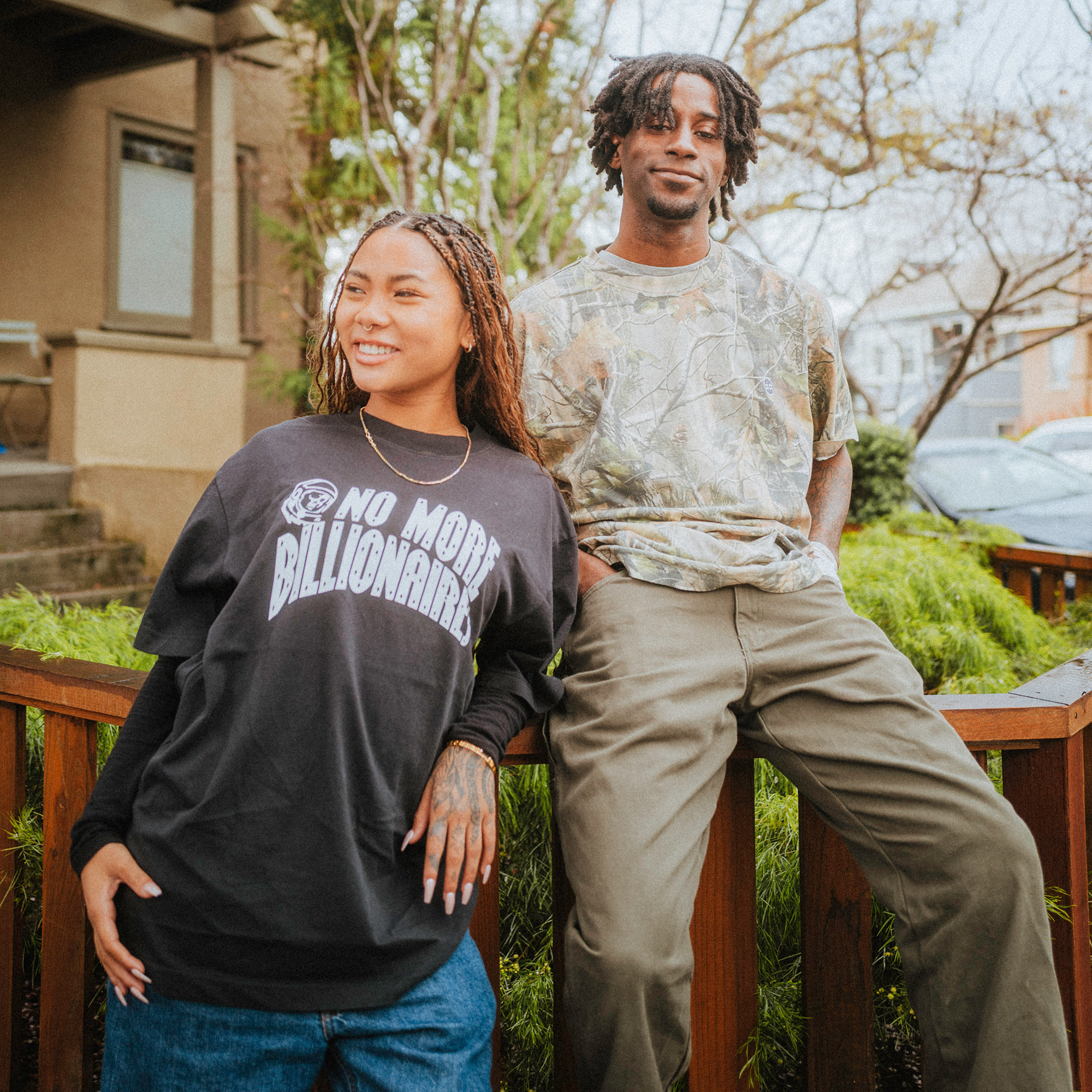 A young woman and man smile outdoors. She wears a BOSK No Billionaires Tee, denim jeans, and her braided hair catches the light. He sports a classic-fit camo shirt with khaki pants as they lean against a wooden railing with greenery behind them.