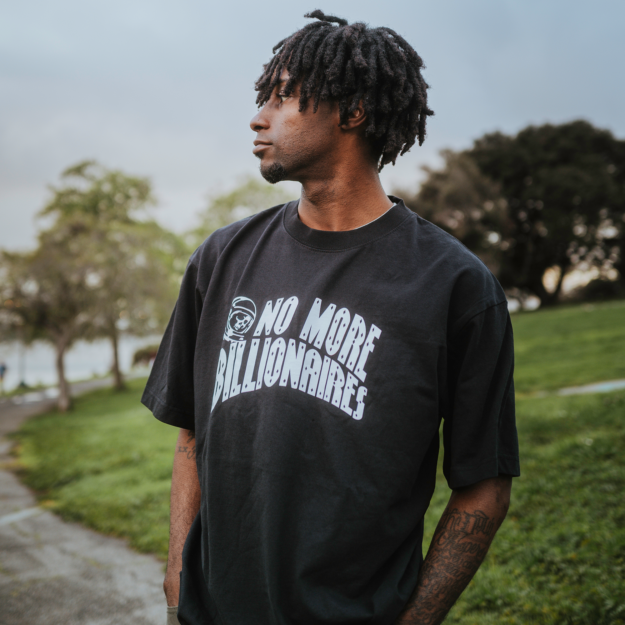 In a park backdrop of green trees and a partly cloudy sky, a man with short dreadlocks gazes to his right while wearing the BOSK "No Billionaires Tee," a classic fit, heavy-weight cotton T-shirt featuring "No More Billionaires" boldly printed in white.
