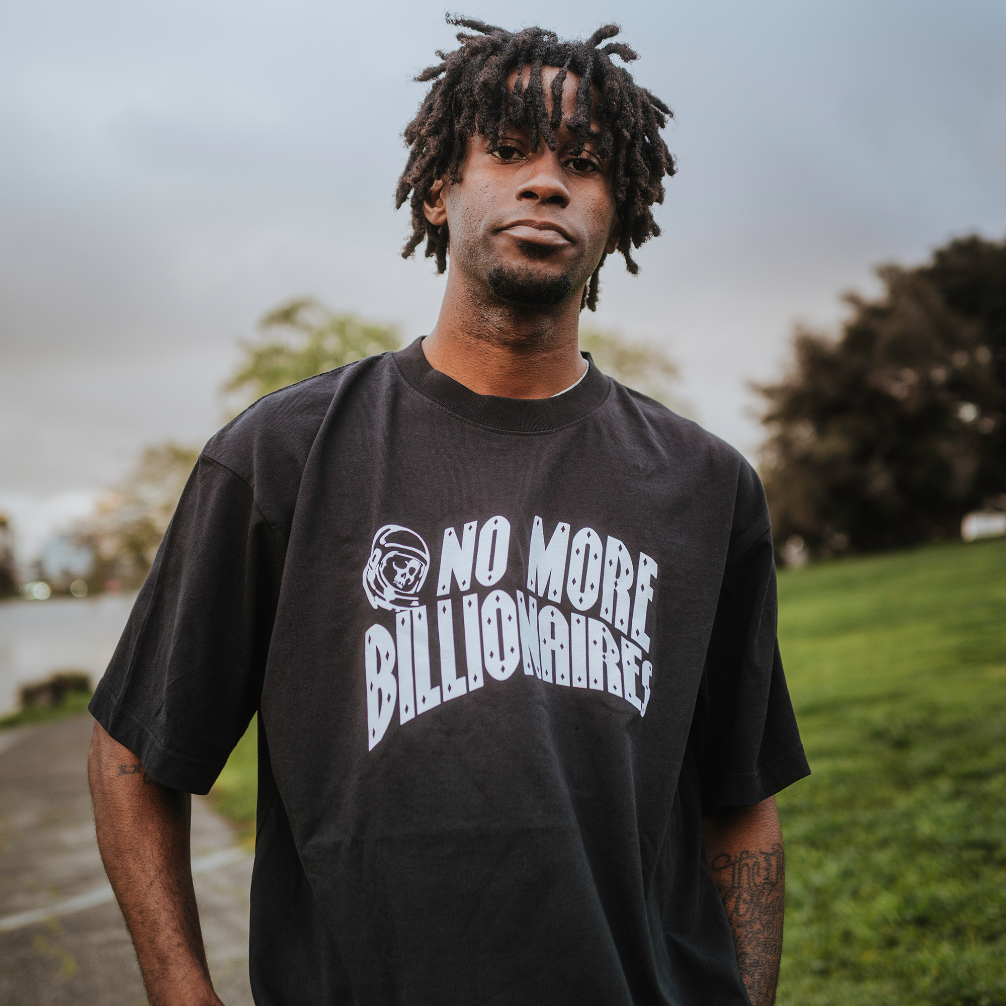 In a tranquil park, a young man with dreadlocks wears BOSK's No Billionaires Tee in classic fit black, featuring "NO MORE BILLIONAIRES" in white. Crafted from heavy-weight 100% cotton, the shirt contrasts beautifully with the green grass, trees, and cloudy sky.