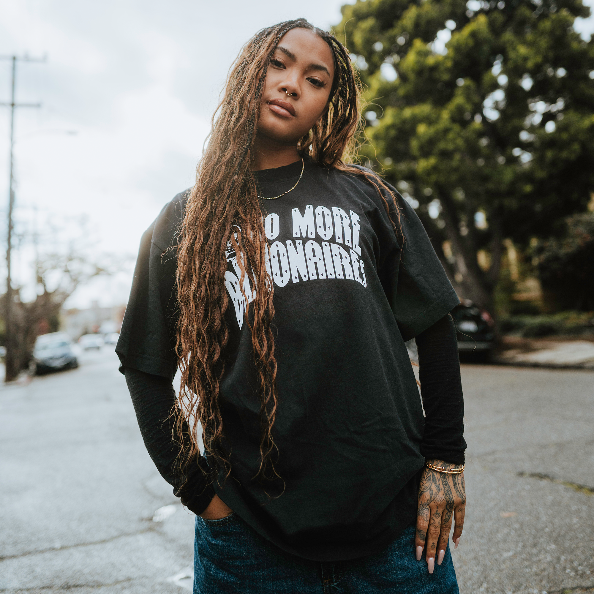 A young person with long braids confidently stands on a quiet street wearing the BOSK No Billionaires Tee, a classic heavy-weight cotton shirt. Under an overcast sky with trees and parked cars in the background, tattoos adorn their hand.