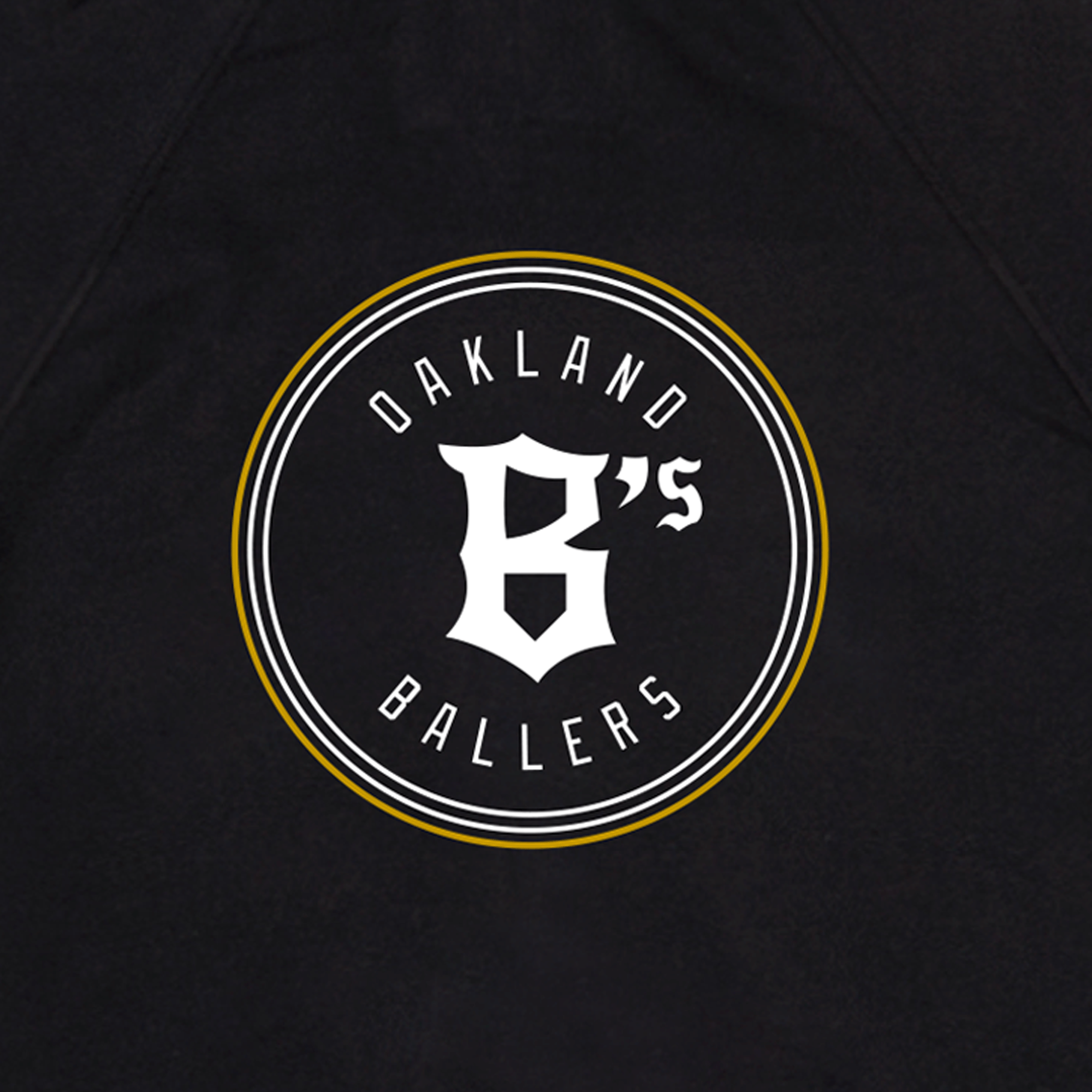 Close-up of round white and gold Oakland Ballers logo and wordmark on the front chest of a black zip up hooded sweatshirt.