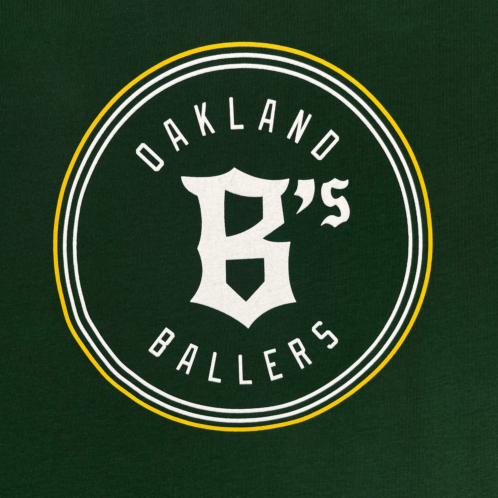 The Women's Oakland Ballers Logo Tee from the Oakland Ballers features a green background with a circular logo that has double white and yellow borders. Inside the circle, "Oakland Ballers" is written in white capital letters encircling a stylized white "B's" in the center, proudly representing the Oakland baseball team competing in the Pioneer League.