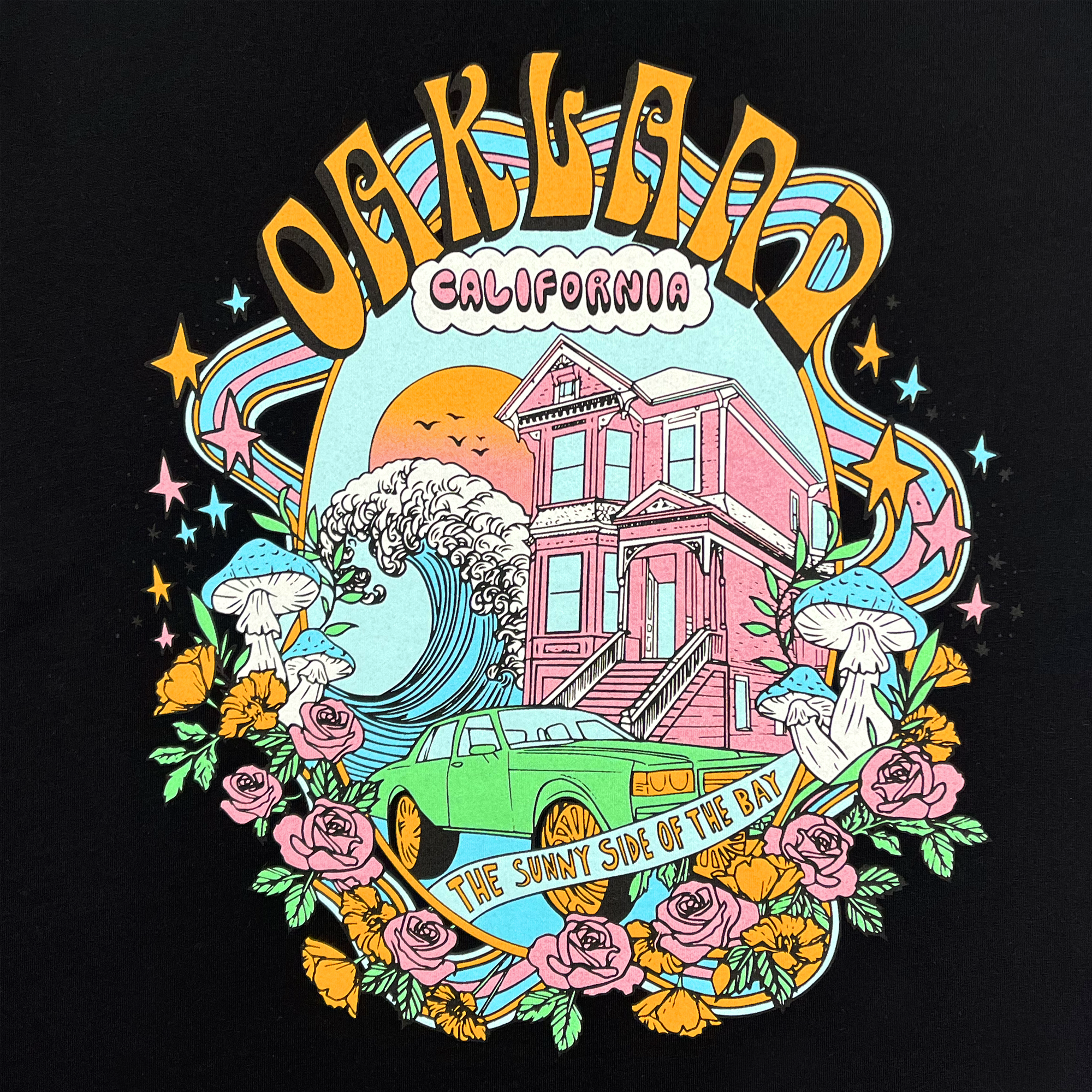 The Women's Oakland Dream Tee from Oaklandish showcases a vibrant graphic on a 100% cotton, classic fit t-shirt. The design features the text "Oakland, California" at the top, displaying a classic Victorian house, a large wave, a green car, mushrooms, roses, and the tagline "The Sunny Side of the Bay" on a banner. The black base is adorned with colorful swirls, stars, and a setting sun in the background.