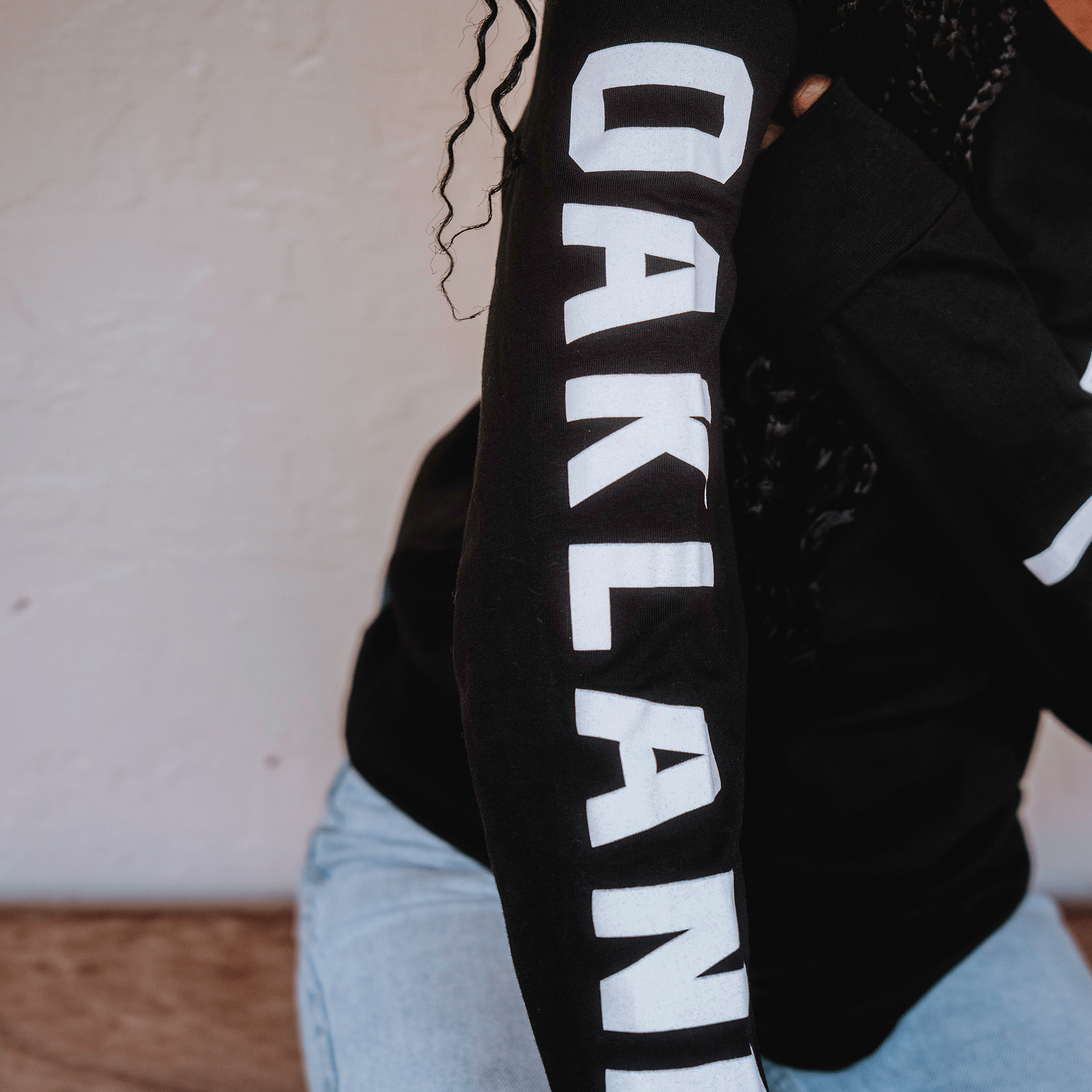 A person wears light blue jeans and a black Oakland Roots SC Classic 2.0 LS Tee with "OAKLAND" in bold white letters down the right sleeve. Their face is unseen as they sit against a textured light wall, embodying community spirit.