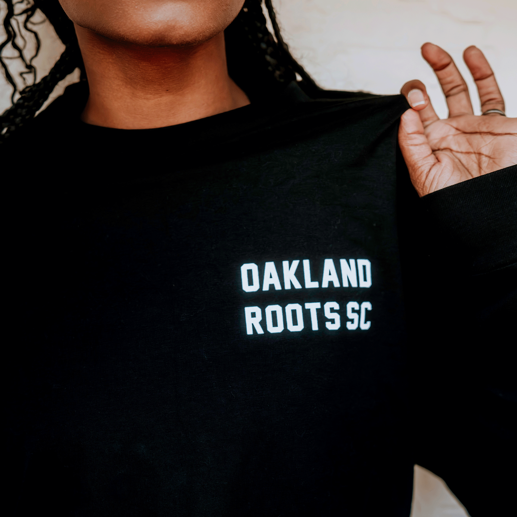 A person with medium brown skin and braided hair wears the Oakland Roots SC Classic 2.0 LS Tee by Oakland Roots SC. Only the lower half of their face and one raised hand are visible against a light background.
