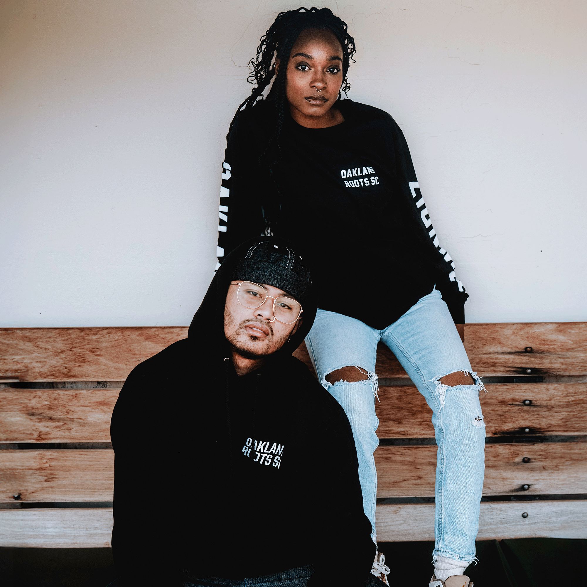 Two people sit on a wooden bench in Oakland Roots SC Classic 2.0 LS Tees, reflecting the club’s commitment to social good and community impact as they enjoy a shared moment against a white wall.