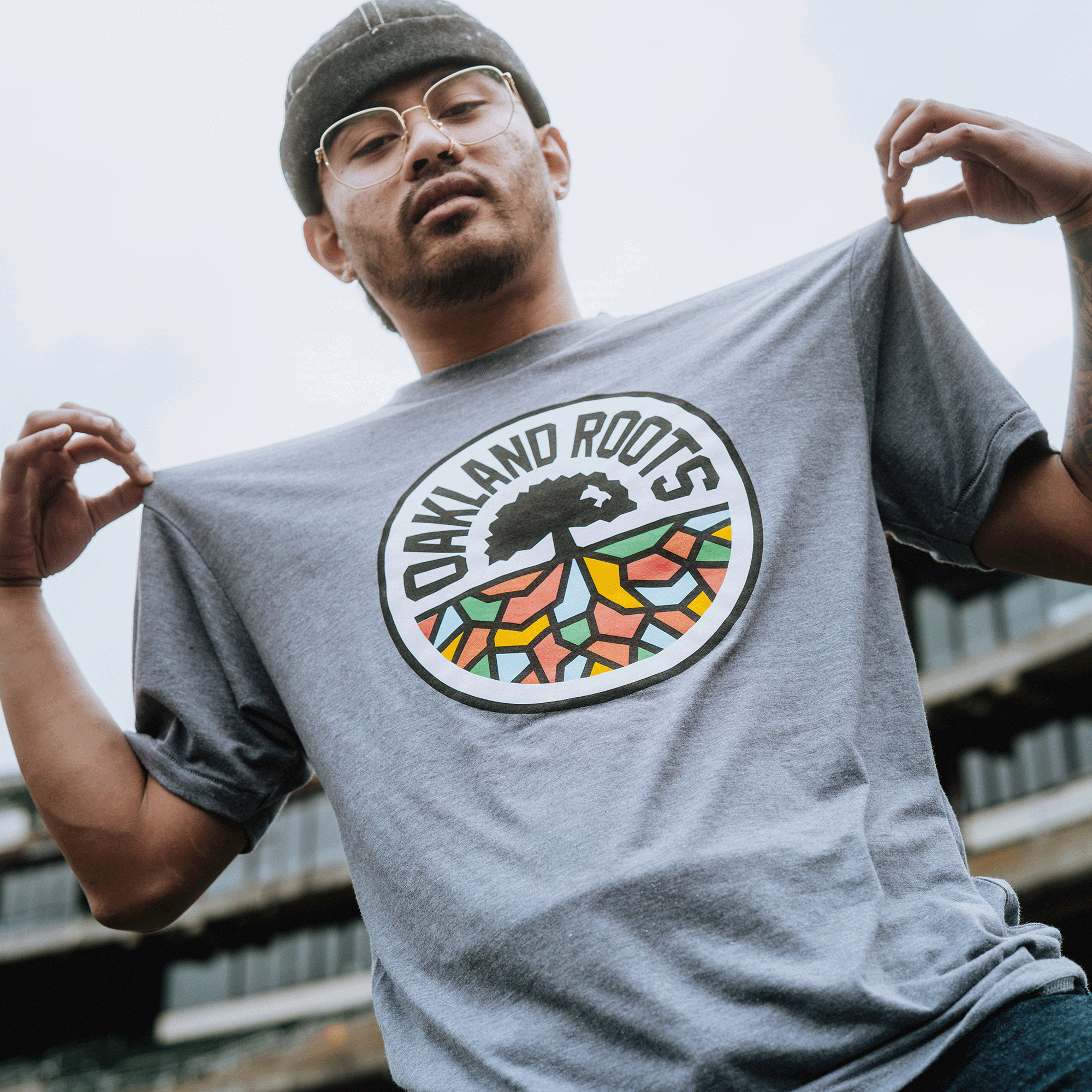A man wearing glasses and a grey beanie displays the Oakland Roots SC Classic Tee by holding out his shirt, showcasing its colorful round "Oakland Roots Sports Club" logo with tree and roots. He stands outdoors, blurred buildings in the background.