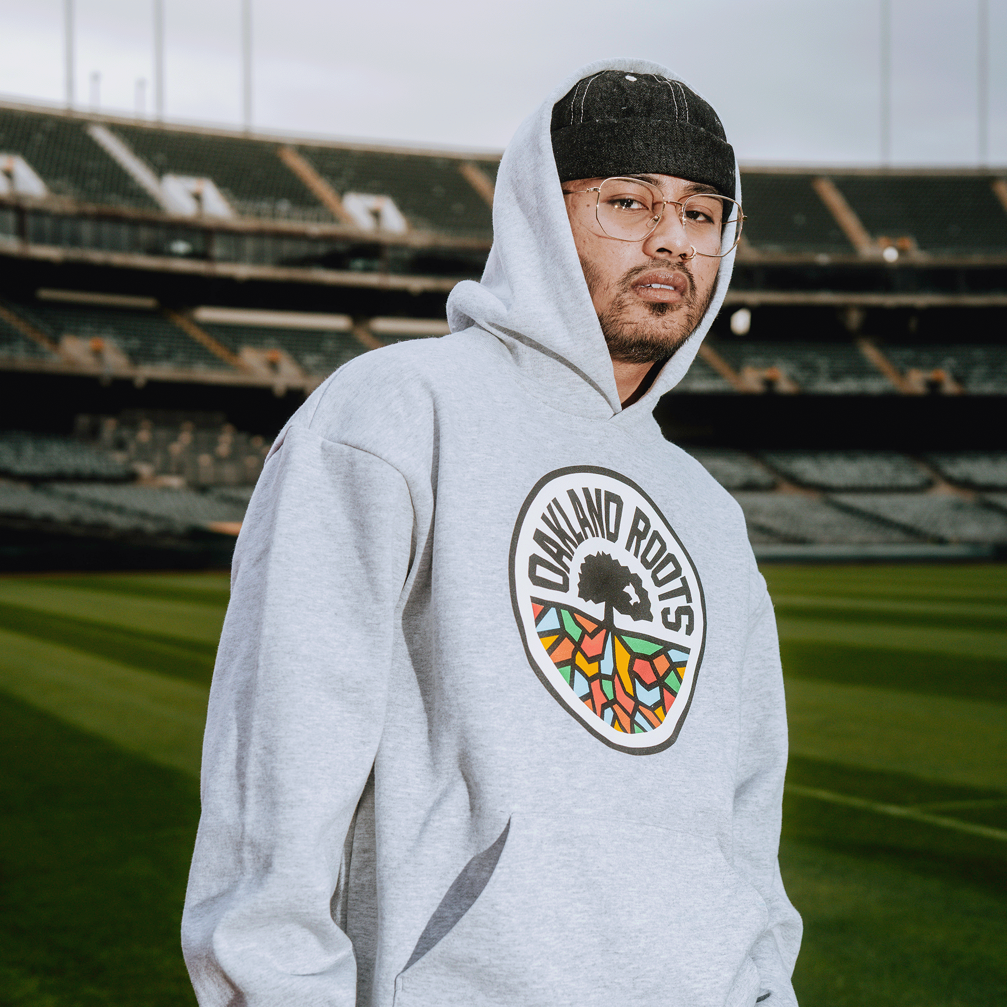 A person stands on a stadium field in an Oakland Roots SC Heavy Hoodie by Oakland Roots SC, wearing glasses and a dark cap. The empty sports club stands and cloudy sky suggest it’s an overcast day.