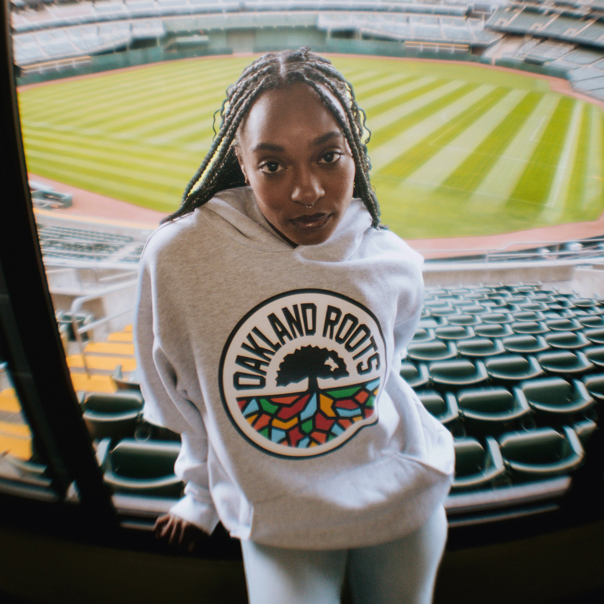 A woman with braided hair wears the Oakland Roots SC Heavy Hoodie from Oakland Roots SC, standing confidently in front of empty stadium seats with the green, striped baseball field behind her, embodying the team’s spirit.