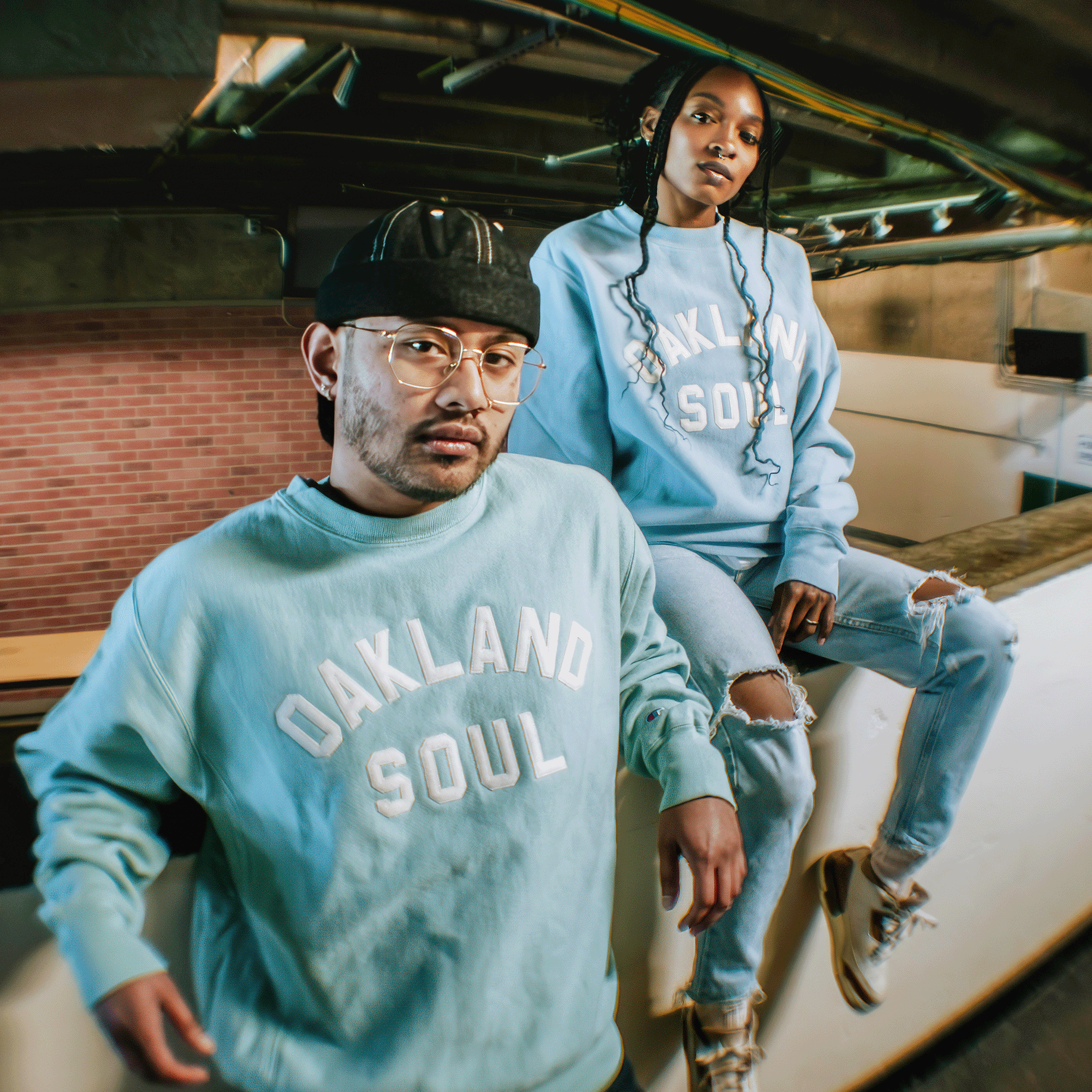 Two people pose indoors in matching Champion X Oakland Soul Wordmark Crew sweatshirts and jeans, showcasing the Oakland Soul brand of the USL W-League. A brick wall and exposed pipes create a laid-back, urban vibe.
