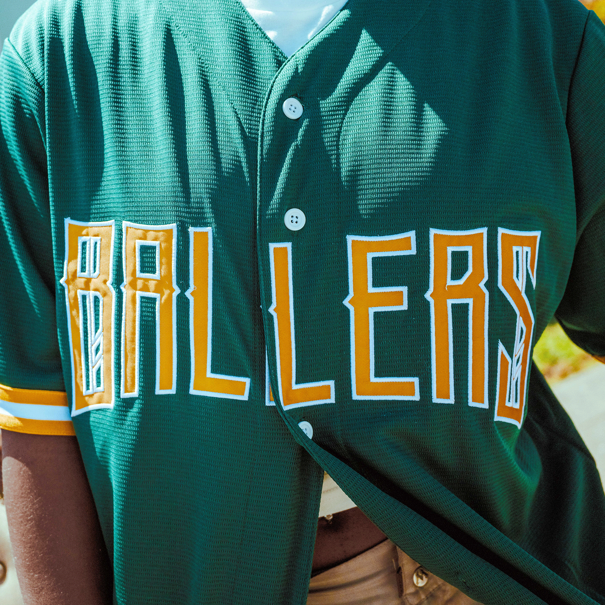 Close-up of a person wearing the Oakland Ballers Button Up Jersey by Oakland Ballers, featuring green fabric with yellow and white trim and "BALLERS" in bold yellow and white letters across the chest, representing the Pioneer League baseball team.