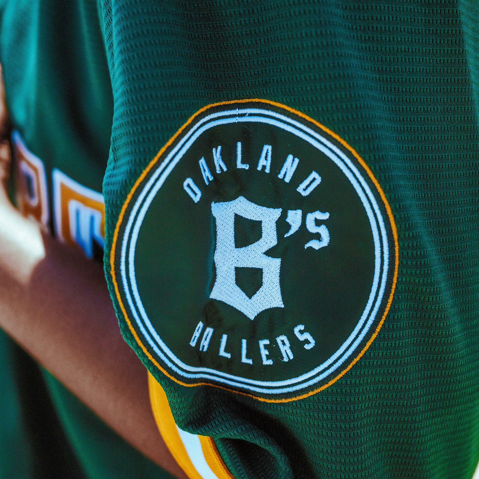 A close-up of the Oakland Ballers Button Up Jersey by Oakland Ballers shows a green sleeve with a circular "Oakland B's Ballers" patch in white, yellow and white borders, and matching stripes near the cuff.