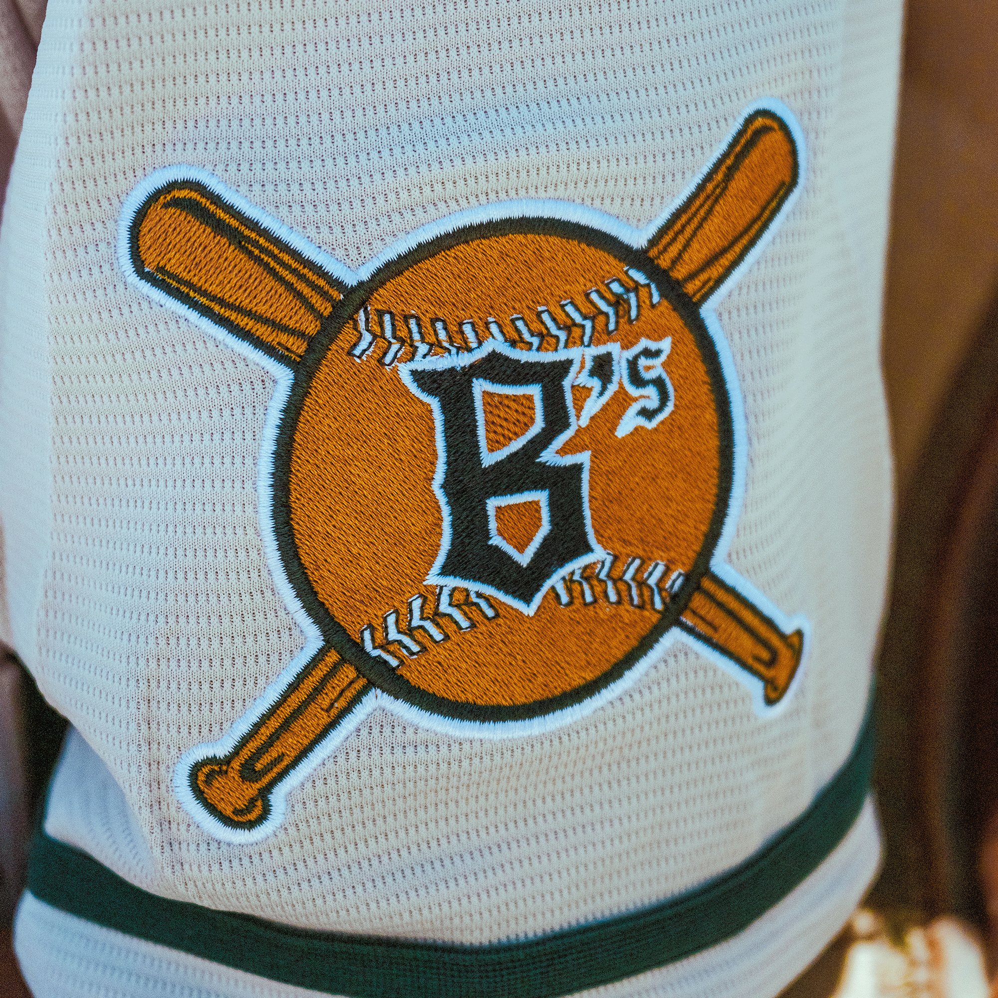 A close-up of the Oakland Ballers Button Up Jersey by Oakland Ballers shows a white fabric patch with a baseball, two crossed bats, bold black "B," and "75" above—all outlined in orange and black—representing the pro baseball team.