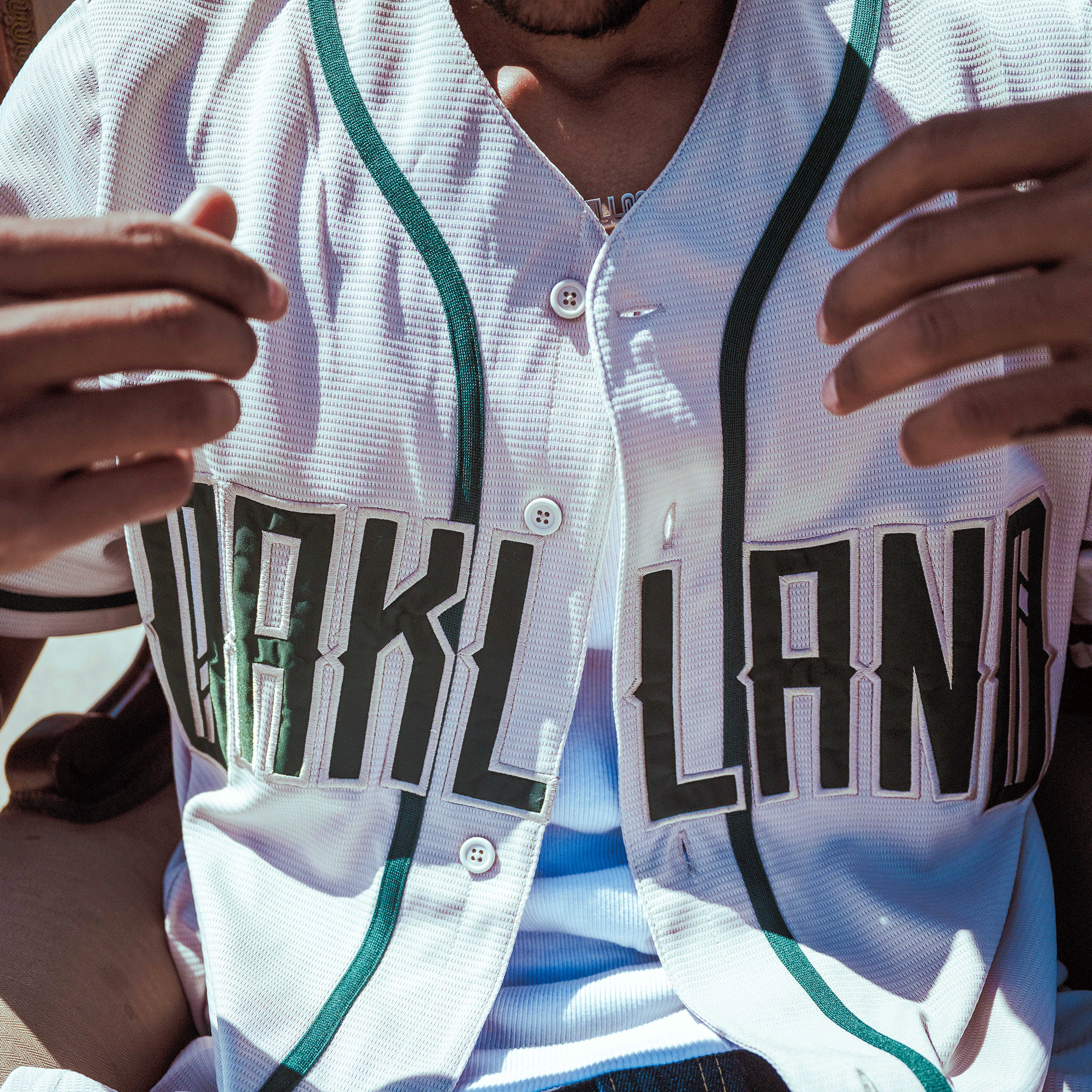 A person wears the Oakland Ballers Button Up Jersey by Oakland Ballers, a white jersey with green trim and bold "OAKLAND" text on the chest. Sunlight creates shadows and blue highlights on the shirt.