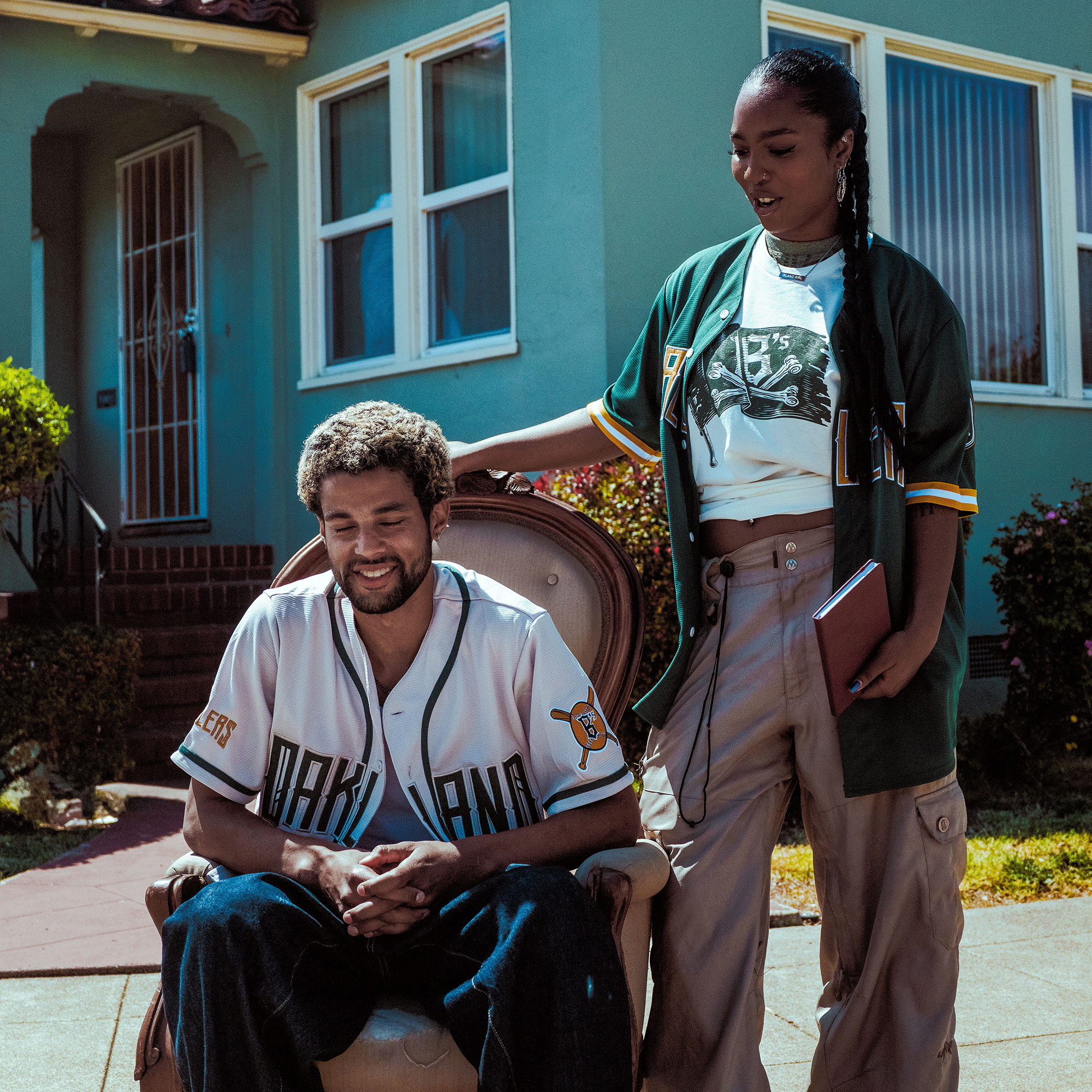 A bearded man with curly hair sits outdoors on an armchair wearing the Oakland Ballers Button Up Jersey, as a woman stands beside him with her hand on his shoulder, both representing their favorite team, the Oakland Ballers, in front of a blue house.