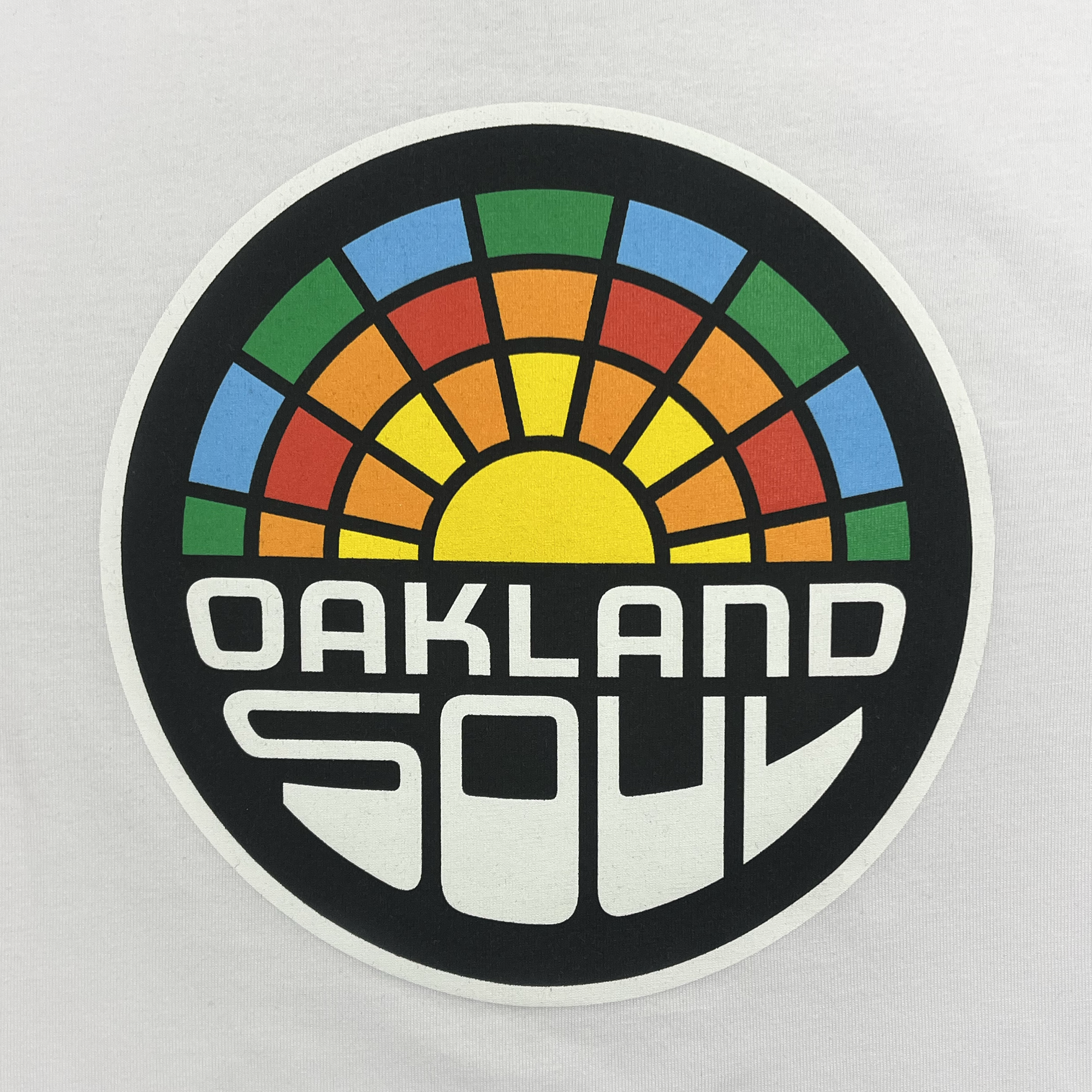 The Youth Oakland Soul Logo Tee features a vibrant circular logo showcasing a stylized sunrise with rays in red, green, orange, blue, and yellow that transition into black. Below the sunrise, "OAKLAND SOUL" is displayed prominently in bold, white geometric typography against the contrasting black background. This dynamic design embodies Oakland Soul's dedication to women's soccer in the USL W-League.