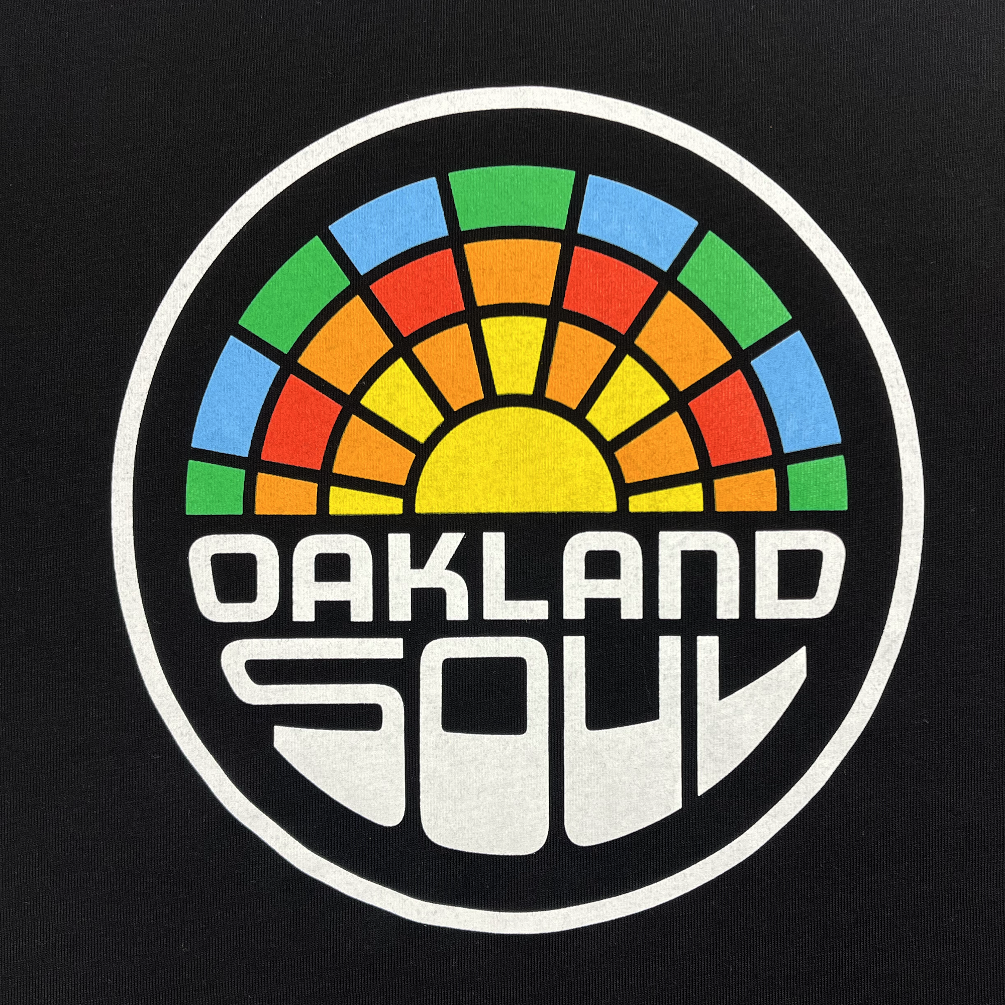 The Infant Oakland Soul Logo One-Piece by Oakland Soul showcases a bold design featuring the words "OAKLAND SOUL" in white, stylized letters at the bottom of a circular logo. Above this text is a vibrant semi-circle resembling a sunset or sun rays with segments in blue, green, yellow, orange, and red. This emblem proudly represents the Oakland Soul women's soccer team in the USL W-League against a sleek black background.