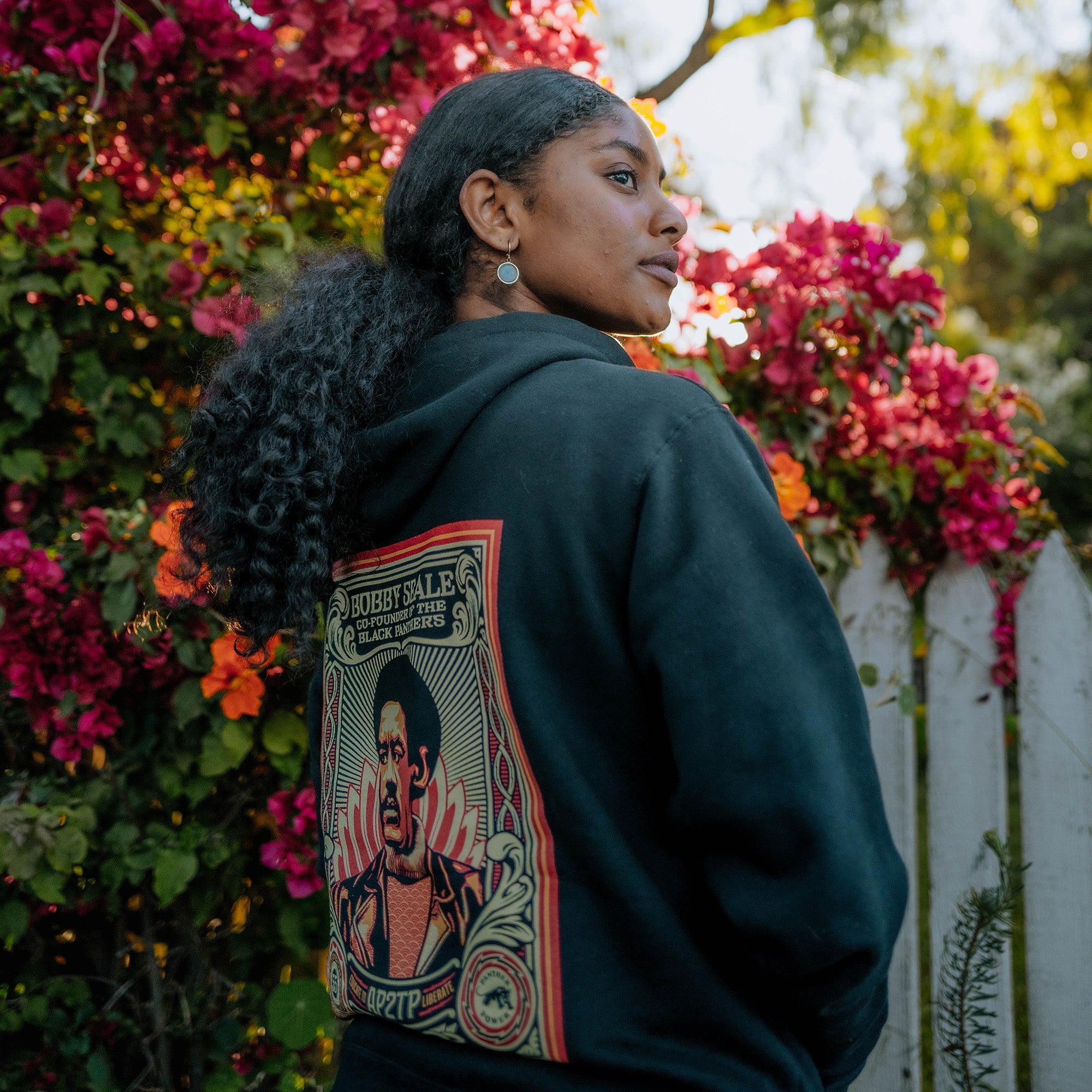 Back angle of female model wearing Educate to Liberate black hoodie outdoors.