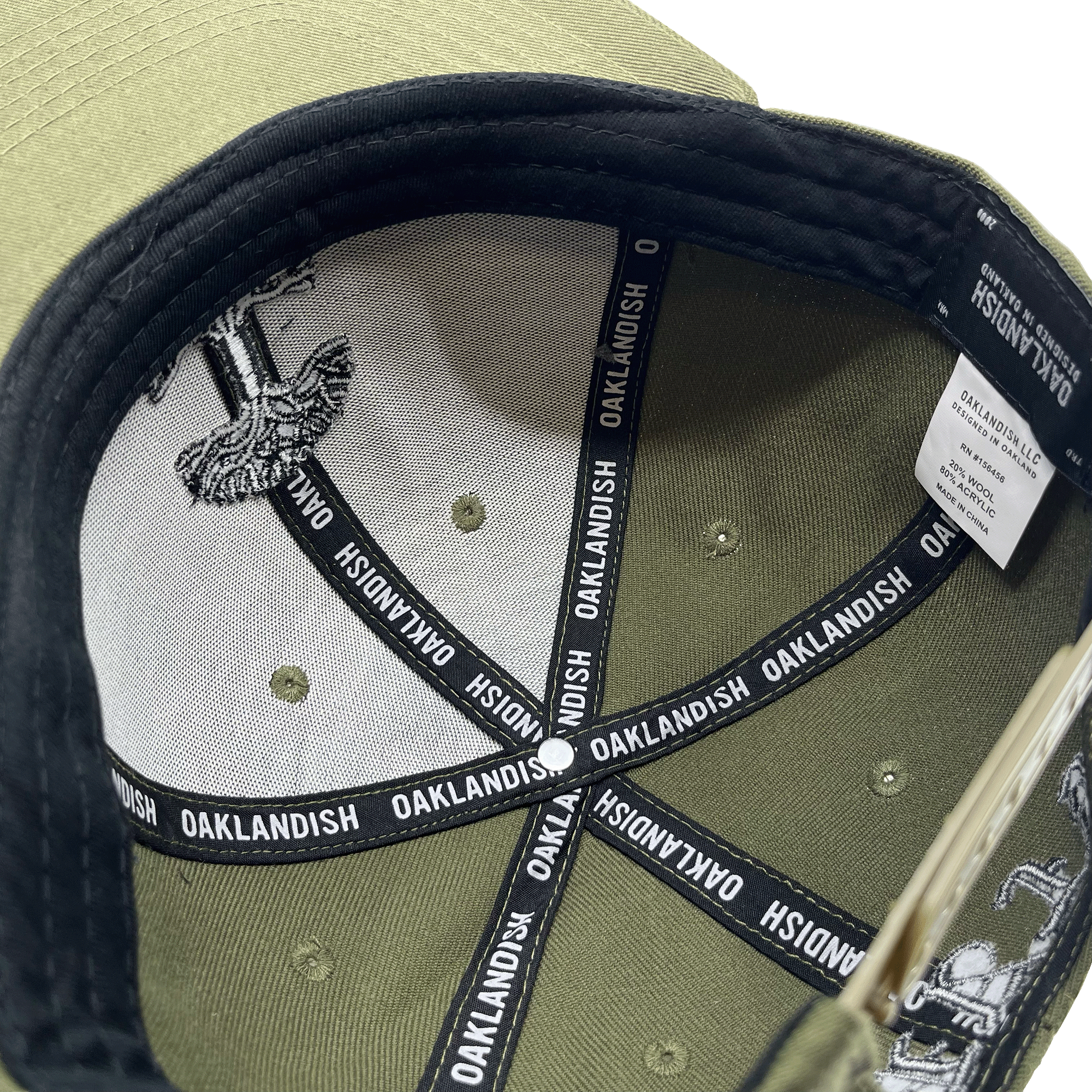 The image showcases the inside of an olive green "Old English Aft Snapback" hat by Oaklandish. The interior features black tape strips with the word "OAKLANDISH" repeatedly printed in white. The underside of the cap's brim displays a gray and white design. Additionally, a white label detailing care instructions is stitched into the lining, complementing its adjustable snapback feature.