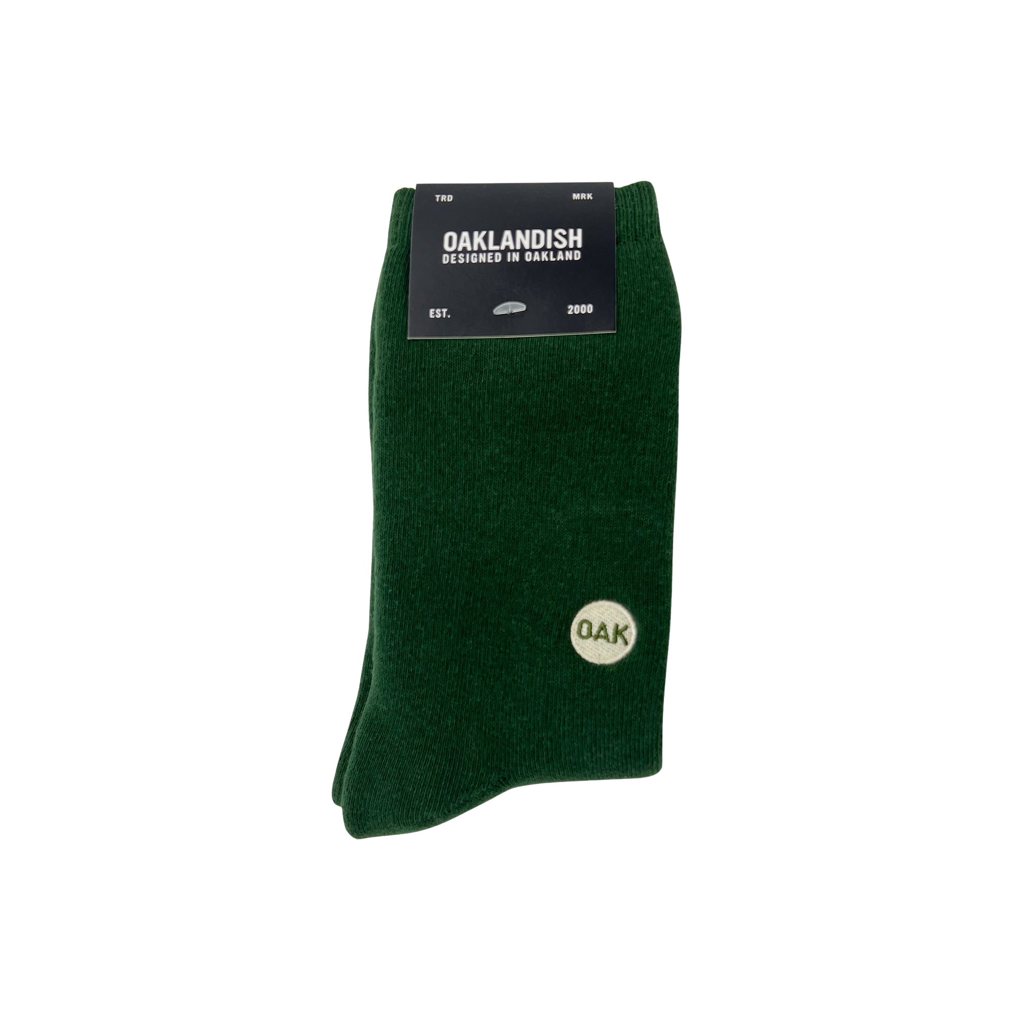 A pair of dark green Plush Oak Socks by Oaklandish are neatly folded and secured with a black label at the top. The label reads “OAKLANDISH,” “Designed in Oakland,” “Est. 2000.” These socks feature a small white circle emblem on the side, containing the letters “OAK” in green.