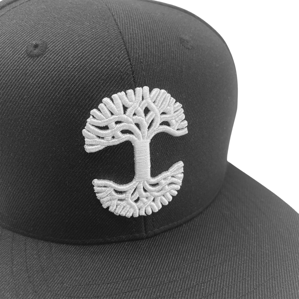 A close-up of the Oaklandish Classic Snapback cap by Oaklandish, highlighting a detailed white embroidered tree logo on its front panel. The design features a symmetrical tree with widespread branches at the top and mirrored roots at the bottom, creating an overall circular pattern. The background is plain white.