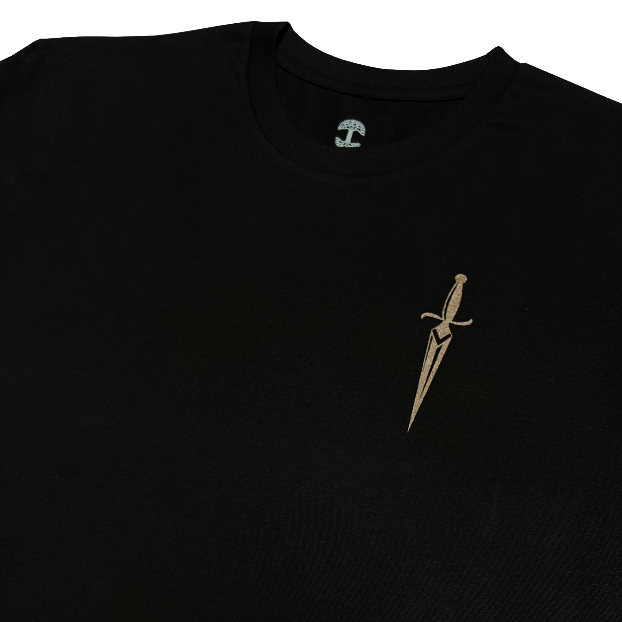 A close-up of the Oaklandish Blossom Tee, a black men's cotton T-shirt featuring a small, intricately designed gold dagger emblem on the upper left chest. The dagger has a sharp, pointed blade and an elegant hilt with stylized details. The plain white background makes the T-shirt and dagger design stand out beautifully.