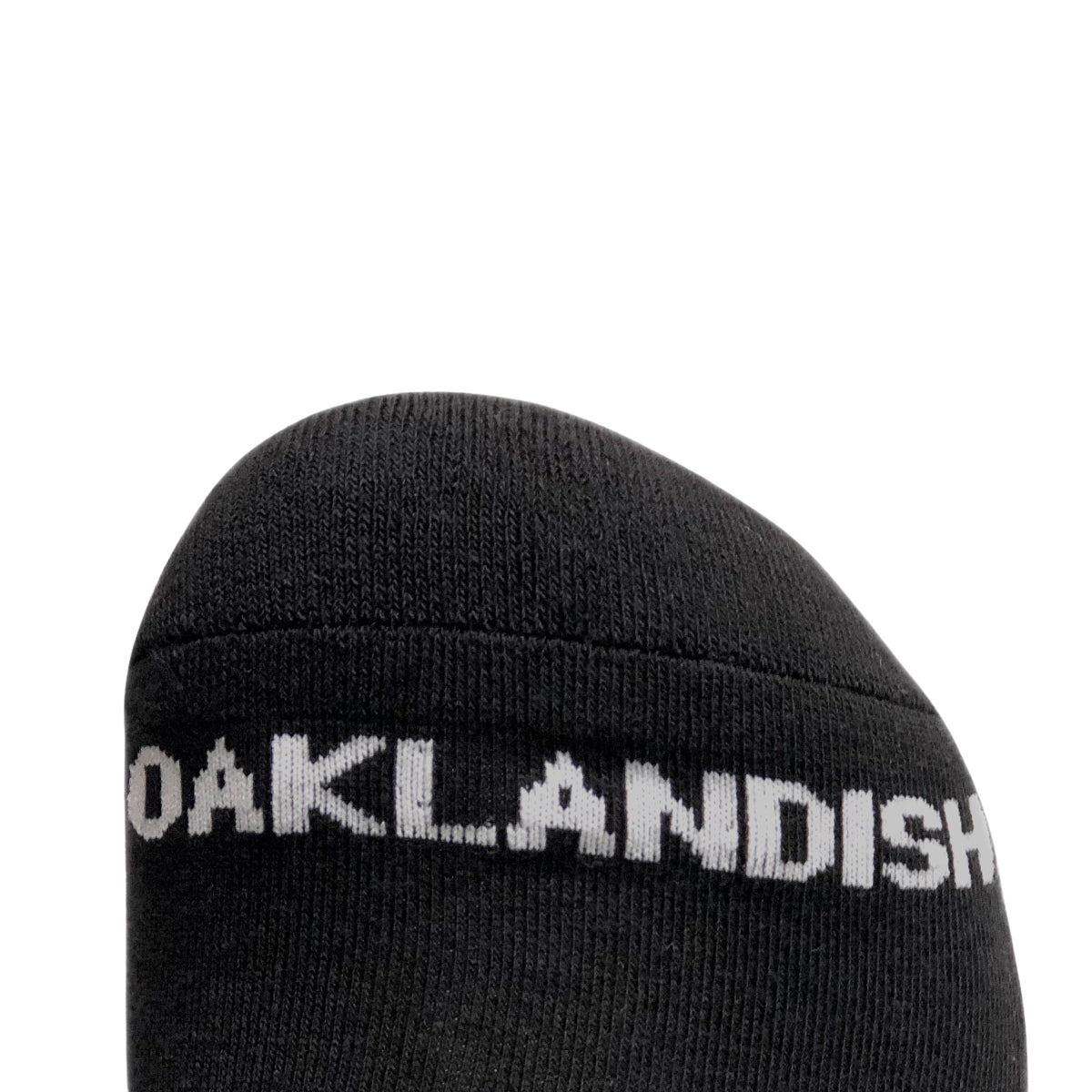 Close-up of the toe section of a black crew sock from the Oaklandish brand's Embroidered Logo Socks collection. The sock features "OAKLANDISH" in white, capital letters, prominently displayed near the front. The background is plain and light-colored, offering high contrast to the black sock and white text. These socks are available in adult sizes.