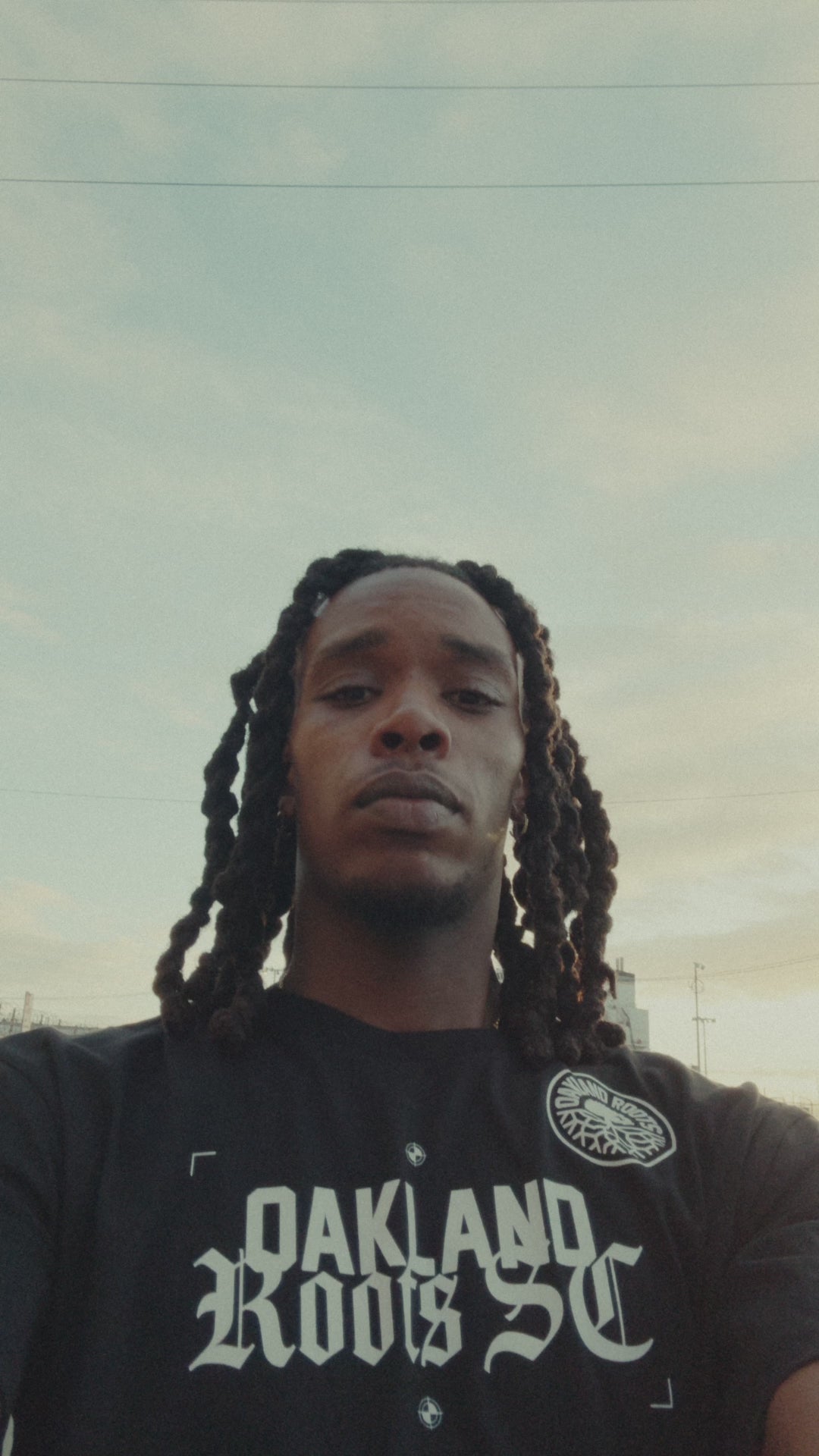 Model walking and skateboarding outdoors in an industrial area of Oakland wearing black tee with Futbol Town design, front and back prints.