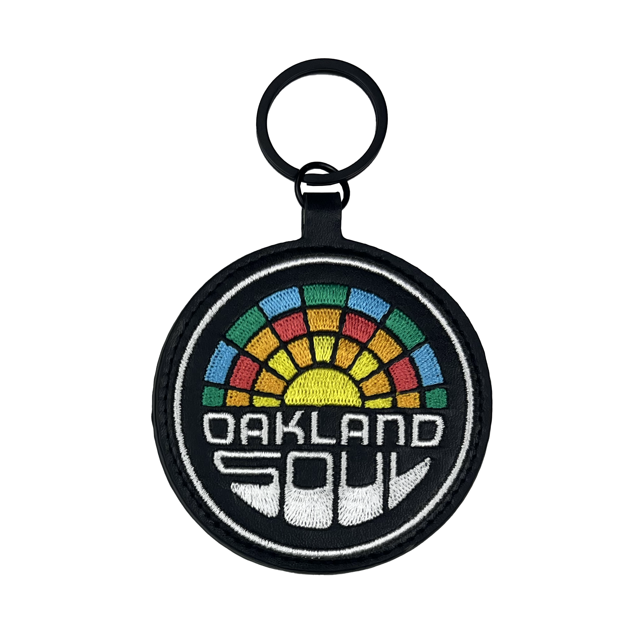 The Oakland Soul Logo Keychain, ideal for fans of Oakland's USL W-League women's soccer team, features a round fabric design with a black border. It showcases the words "OAKLAND SOUL" in white block letters at the bottom, and above this text is a colorful geometric design reminiscent of a rainbow mosaic pattern. The keychain is equipped with a black ring for conveniently attaching keys.