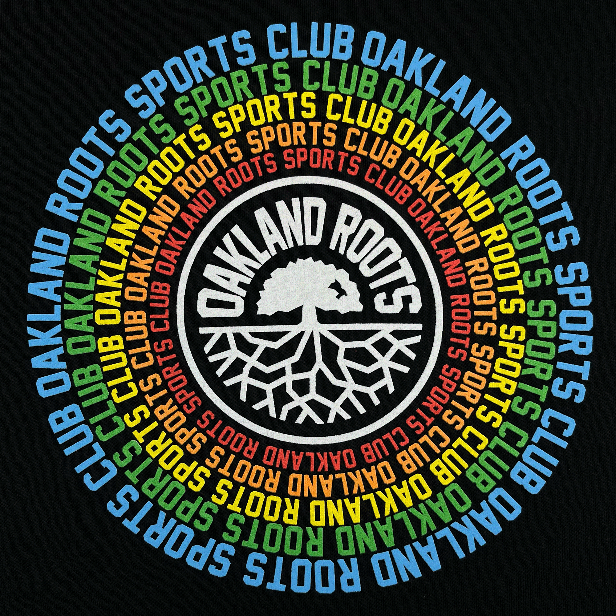 The Oakland Roots SC Aura Tee by Oakland Roots SC features a circular logo with the text "Oakland Roots Sports Club" repeated in a colorful ring of blue, green, yellow, orange, and white. At its center is a white tree with extending roots and the words "Oakland Roots" positioned above the tree image. The black background signifies their commitment to social good.